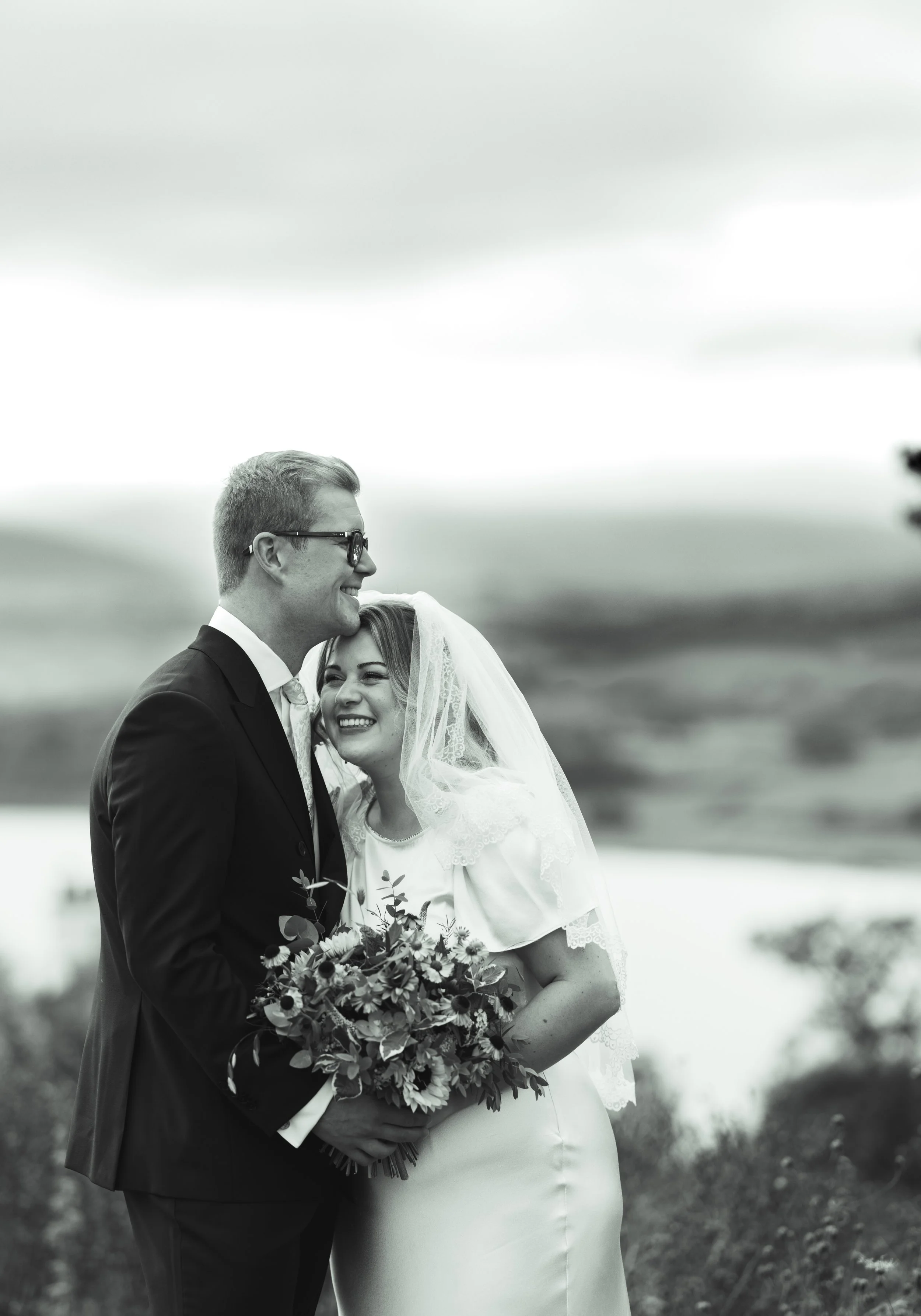 Black and white photograph of a bride and groom on their wedding day, outdoors, smiling and holding a bouquet of flowers, with a blurred landscape in the background.