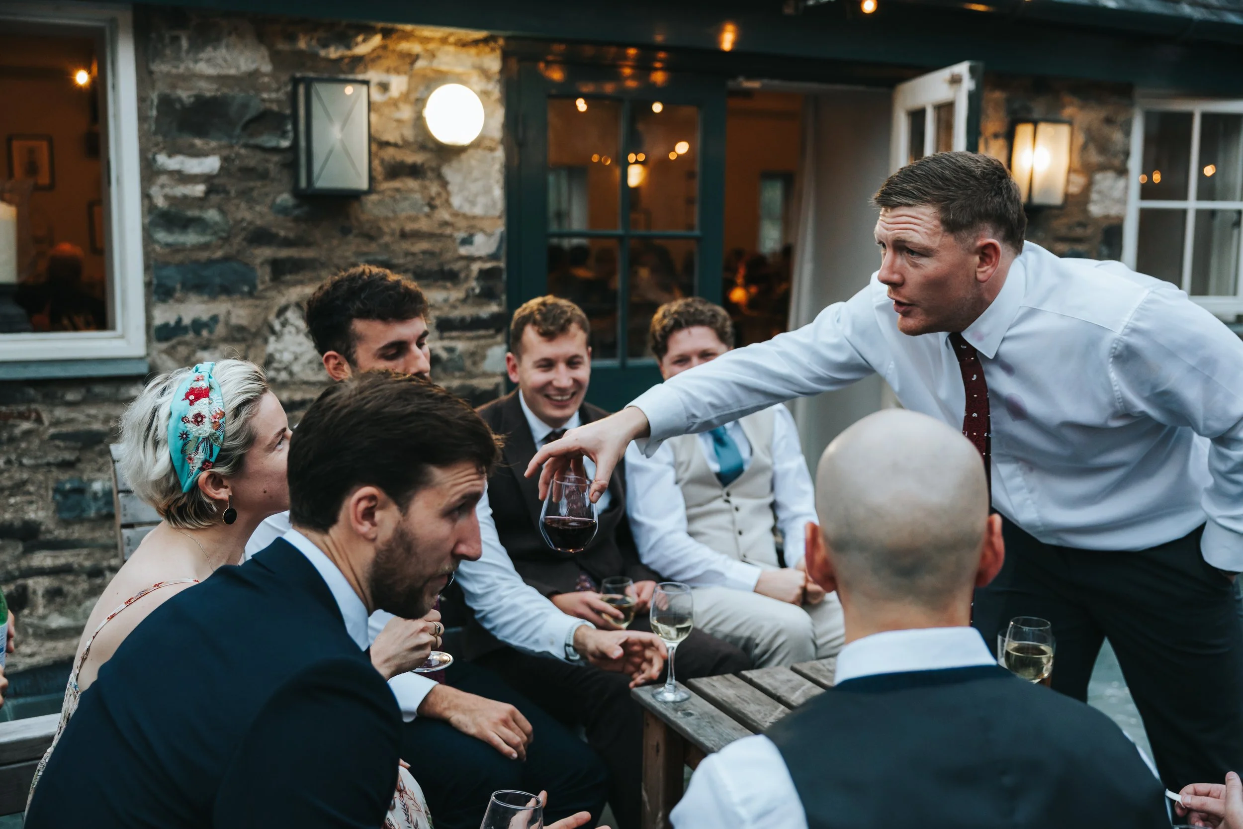 Man in white shirt and red tie pouring red wine into glass in front of group of smiling people at an outdoor gathering