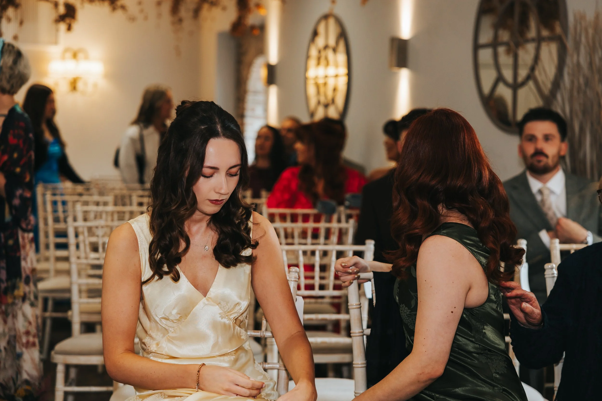 Two women sitting at a social event, one woman with dark, wavy hair in a pale yellow silk dress looking down, while the other woman with red, wavy hair in a black dress adjusts the woman’s sleeve in a well-lit room with wooden chairs and large round 