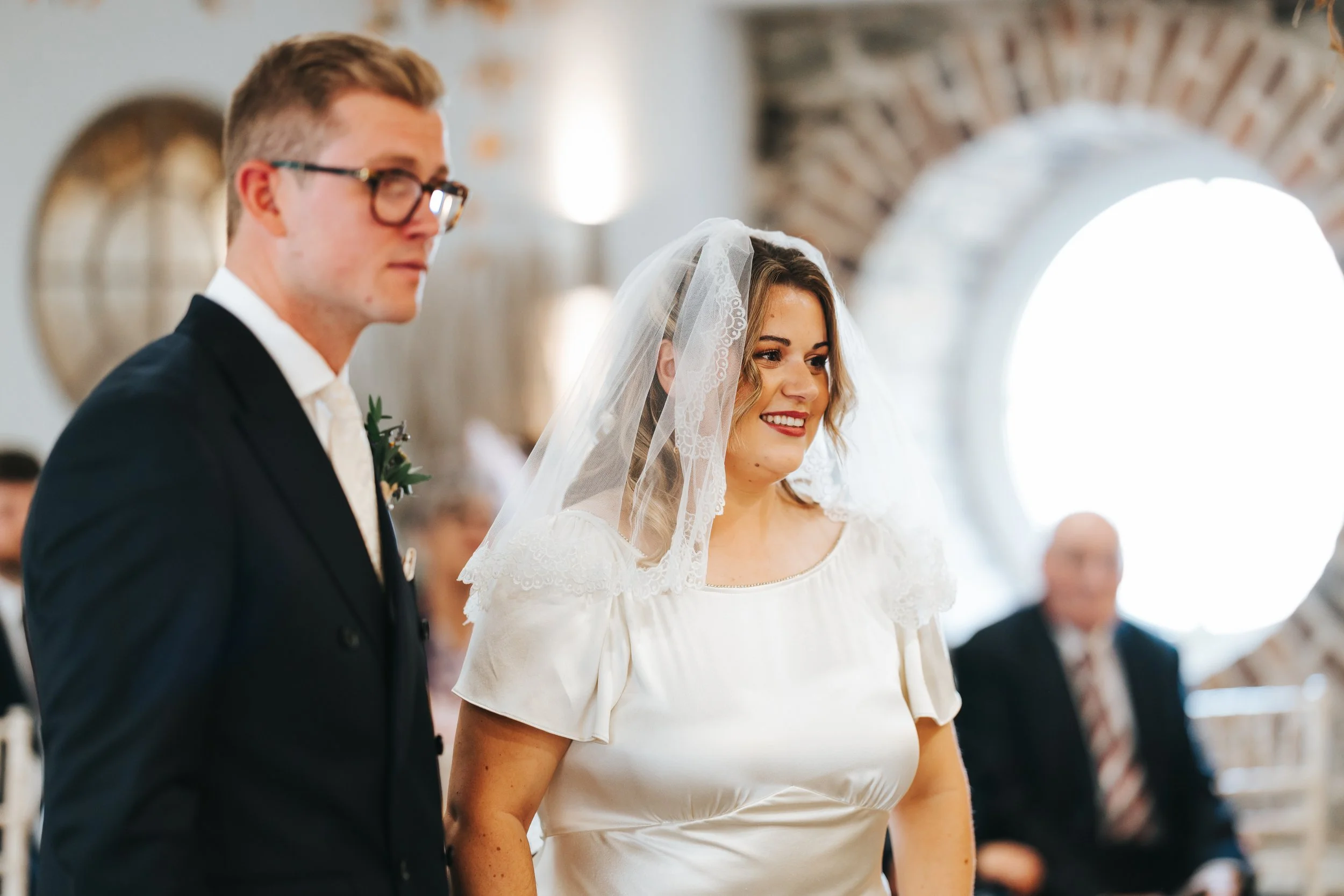 Bride and groom standing during wedding ceremony inside a venue with arched brick wall and round windows, bride smiling and wearing a satin white dress with lace veil, groom in black suit and glasses.