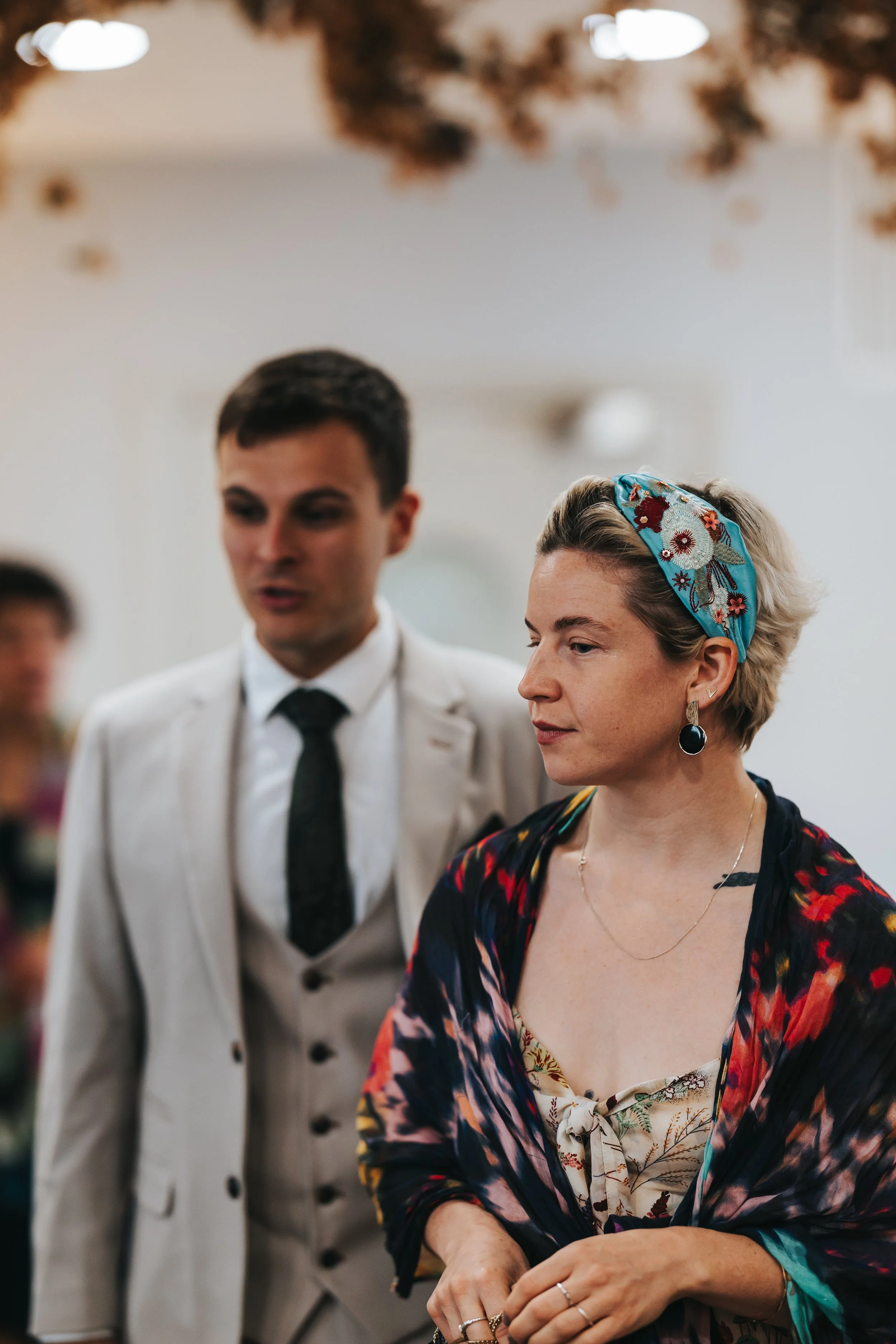 A woman with short blonde hair, wearing a blue floral headscarf, earrings, and a colorful shawl, looking contemplative. A man in a light-colored suit and dark tie stands behind her, possibly at a social gathering or event.