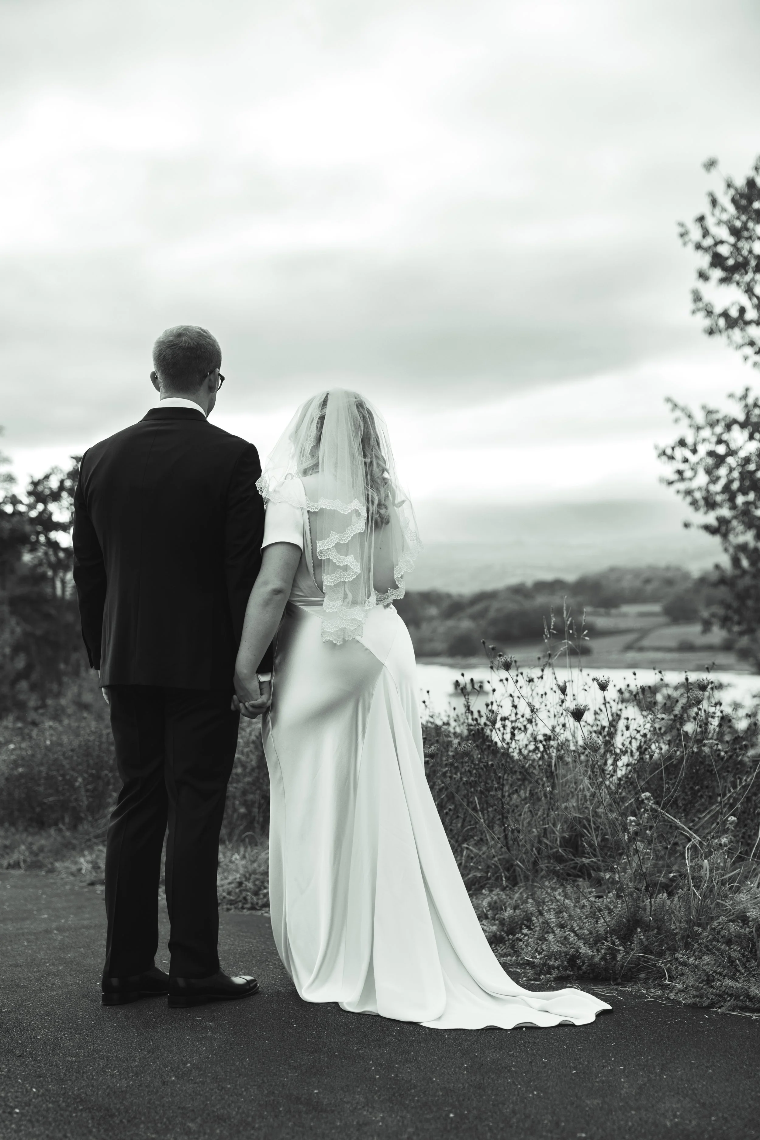 Black and white photo of a bride and groom holding hands, standing outdoors on a cloudy day by a body of water with trees in the background.