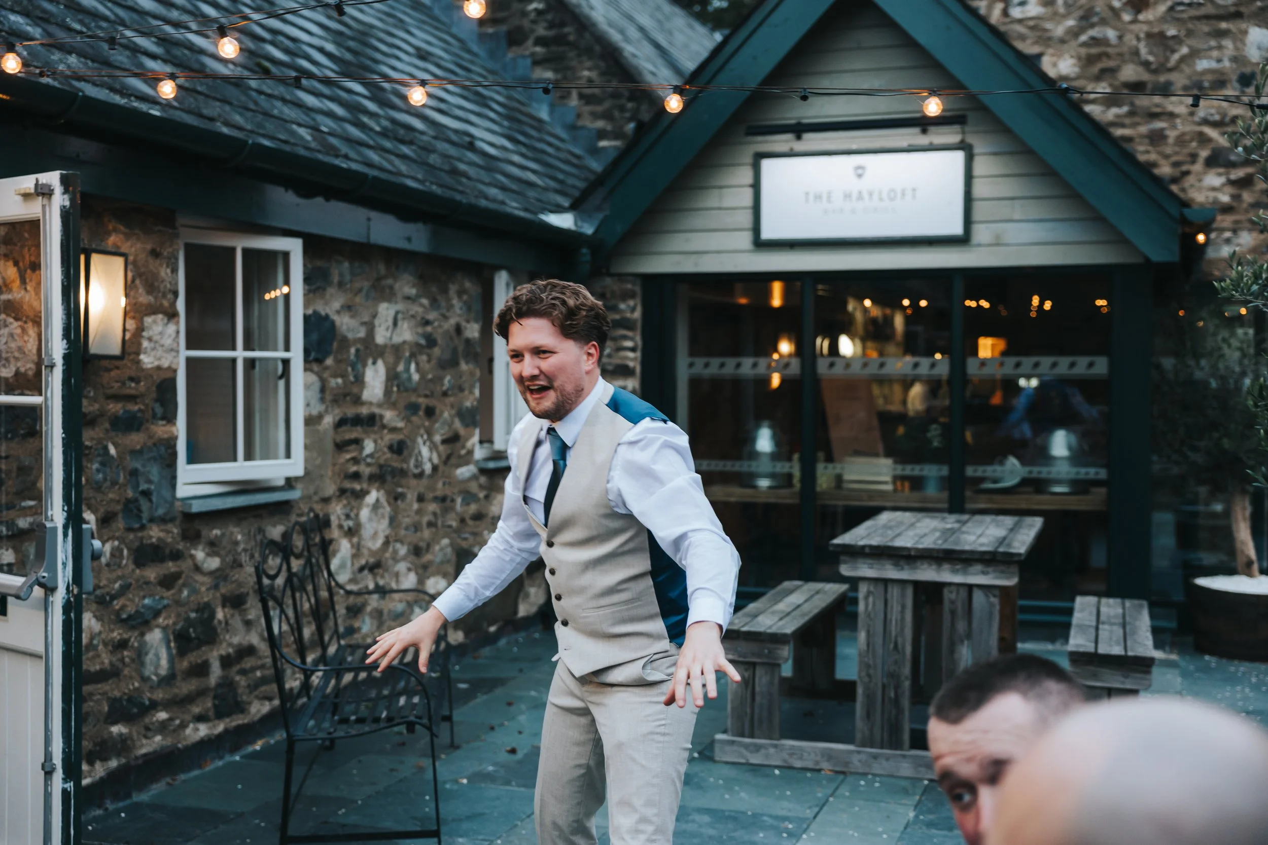 A man in a white shirt, grey vest, and tie, standing outdoors in front of a rustic stone building with string lights, appears to be dancing or acting playfully.