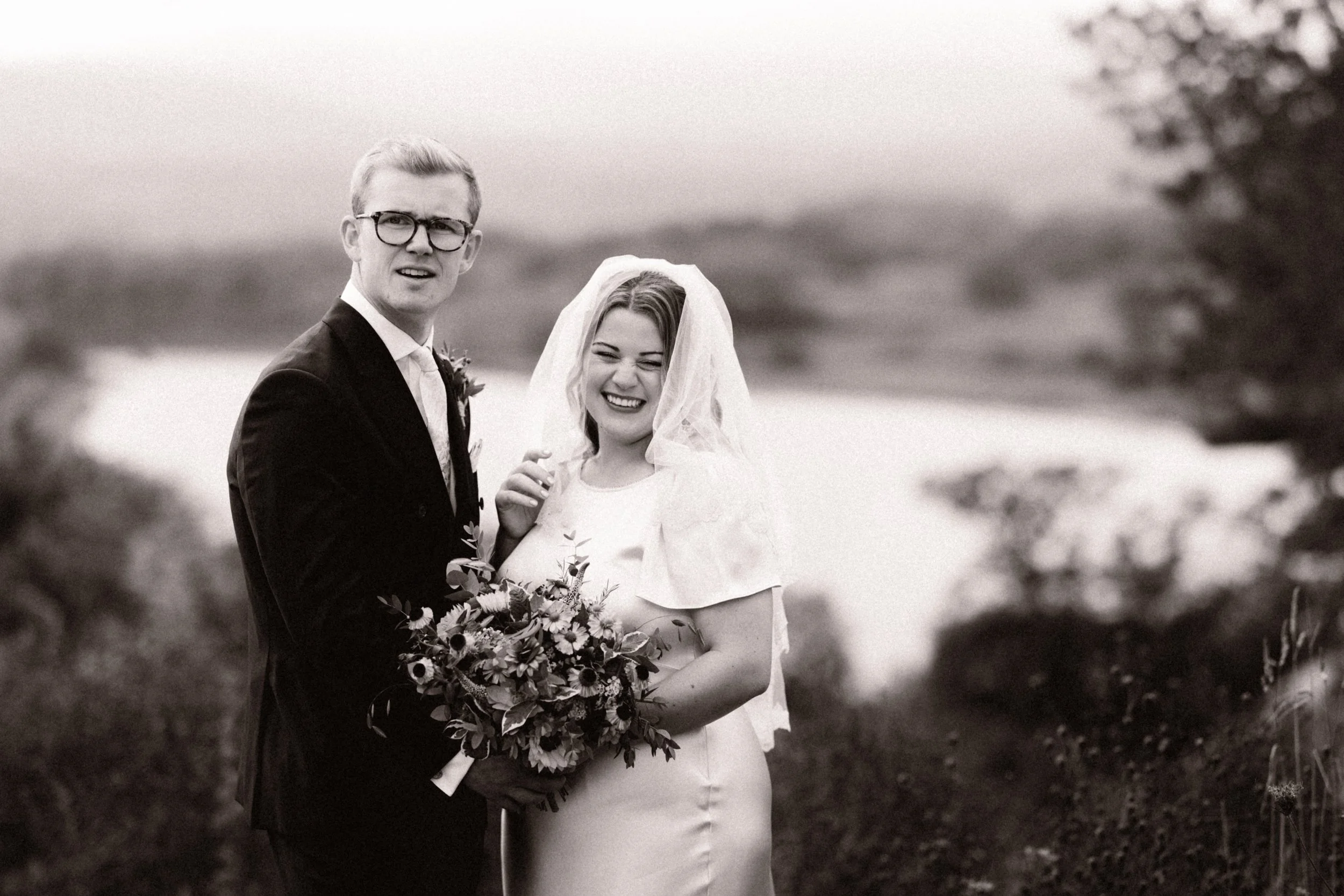 Black and white photo of a groom and bride outdoors, smiling, with the groom in a suit and glasses holding a bouquet, and the bride wearing a wedding dress and veil, near a body of water and surrounded by trees.
