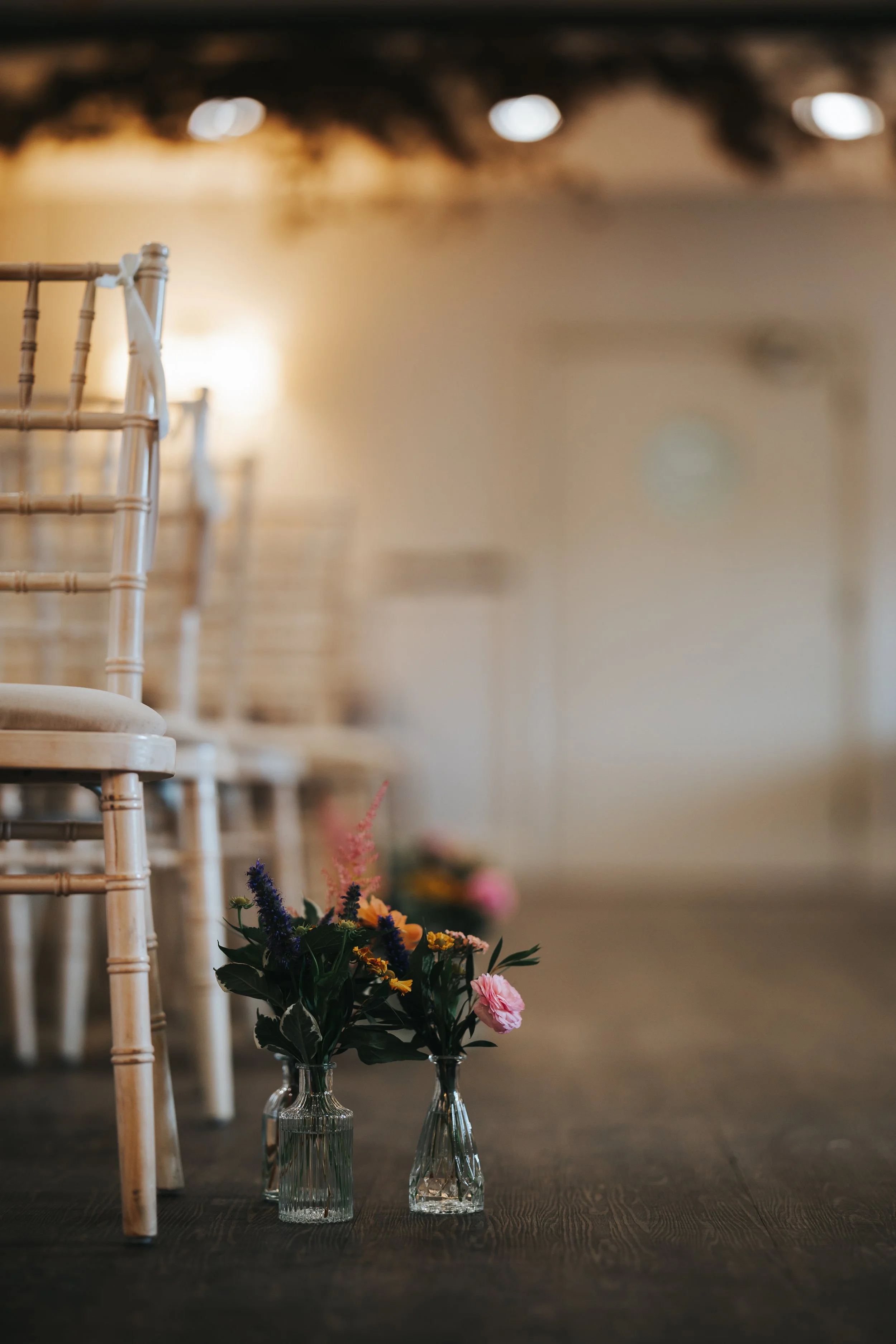 Two small vases with colorful flowers placed on a wooden floor, with chairs arranged in the background.