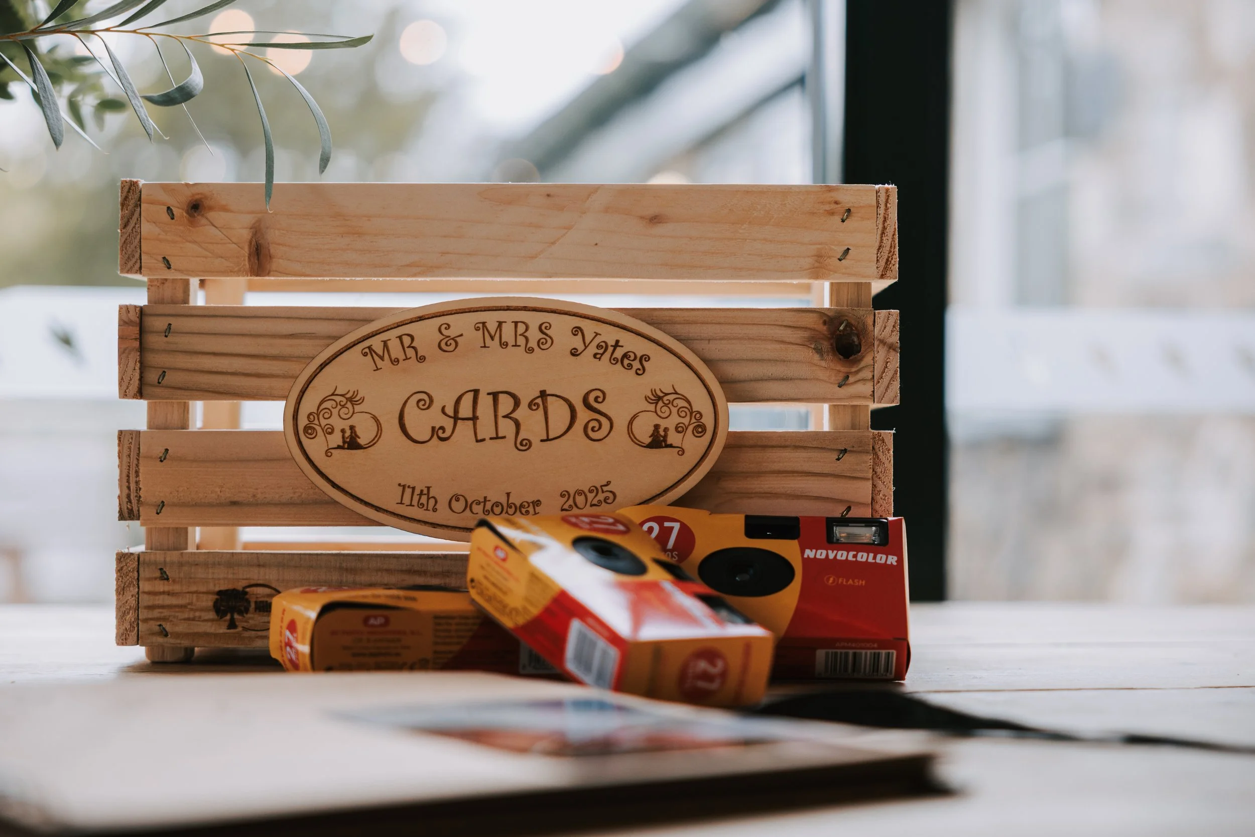 Wooden crate with engraved sign for Mr. & Mrs. Yates Cards, dated October 11th, 2025, on a table with some unopened film rolls in front of it, near a window.