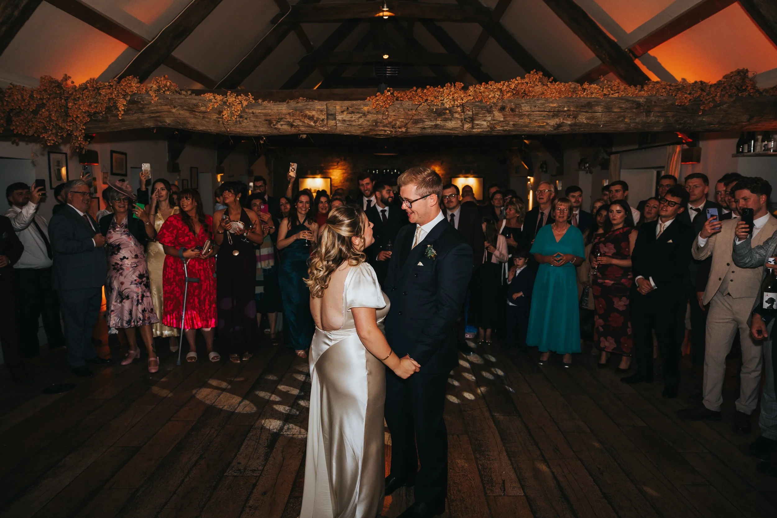 A bride and groom holding hands and dancing at their wedding reception, surrounded by guests in a rustic decorated venue with wooden beams and warm lighting.