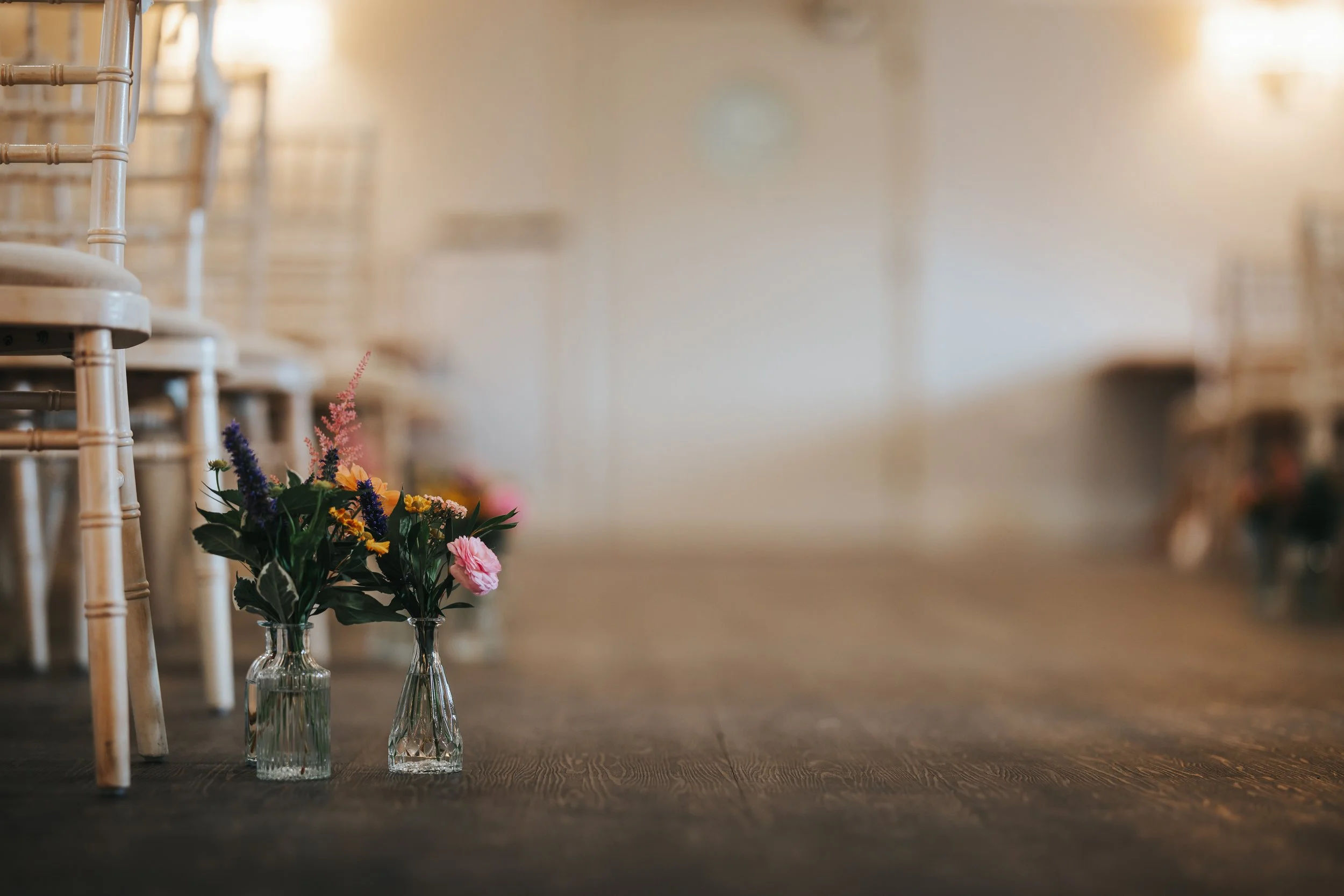 Two glass vases with colorful flowers on a wooden floor, with chairs lined up along the wall in the background.