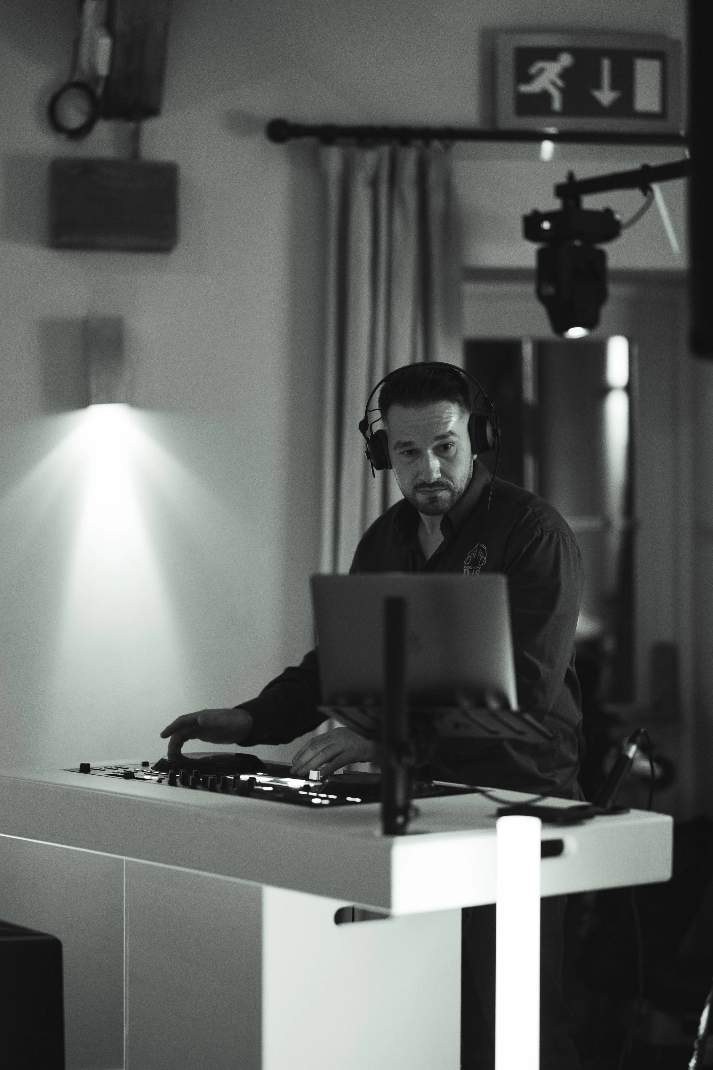 Black and white photo of a DJ wearing headphones, standing behind a DJ mixer and laptop, in a room with curtains and lighting fixtures, with an emergency exit sign overhead.