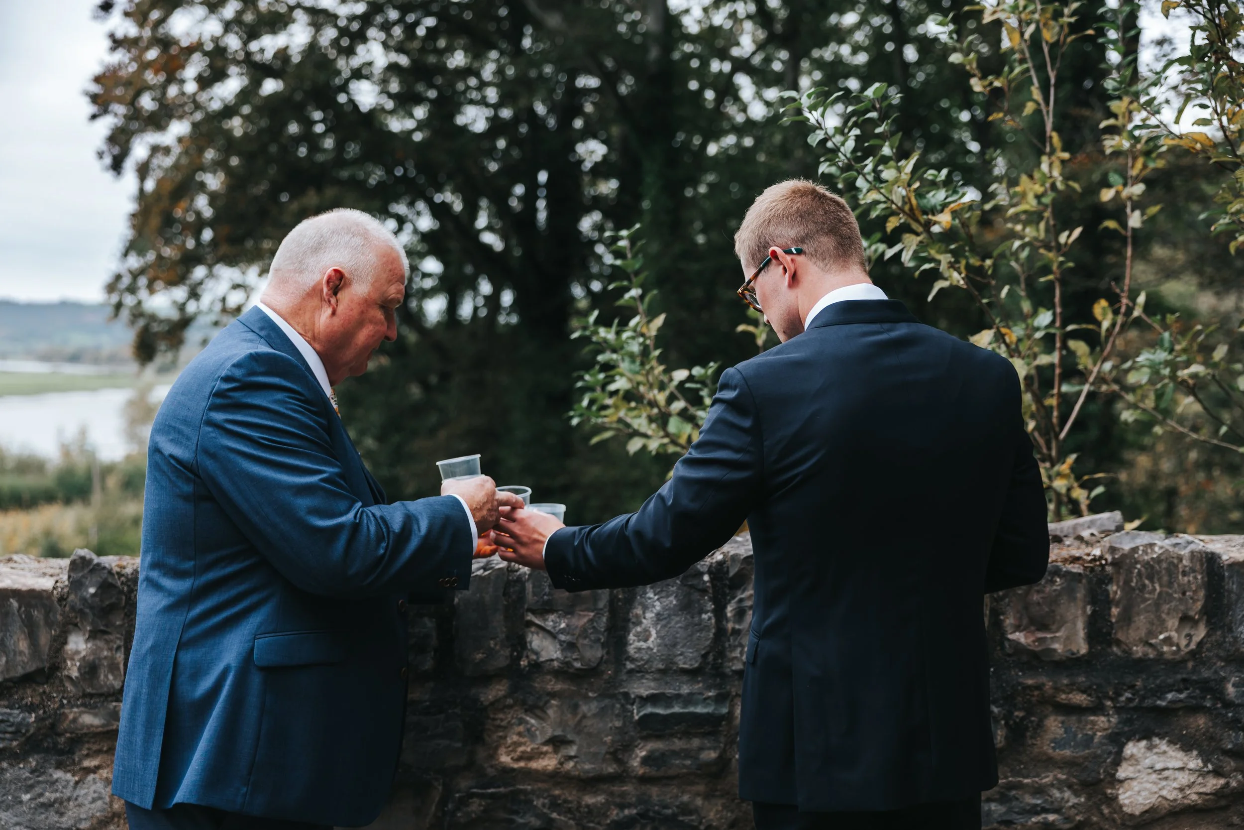 Two men in formal suits pouring drinks into glasses outdoors near a stone wall, with trees and a river in the background.