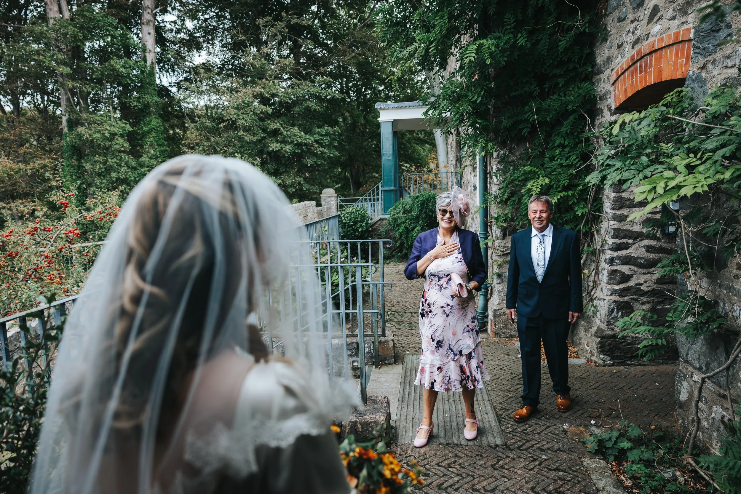 A bride with a veil stands outdoors, facing her parents who are smiling and looking at her. The mother has her hand on her chest, and the father stands beside her, both dressed formally. The scene is on a brick pathway with green foliage and a stone 