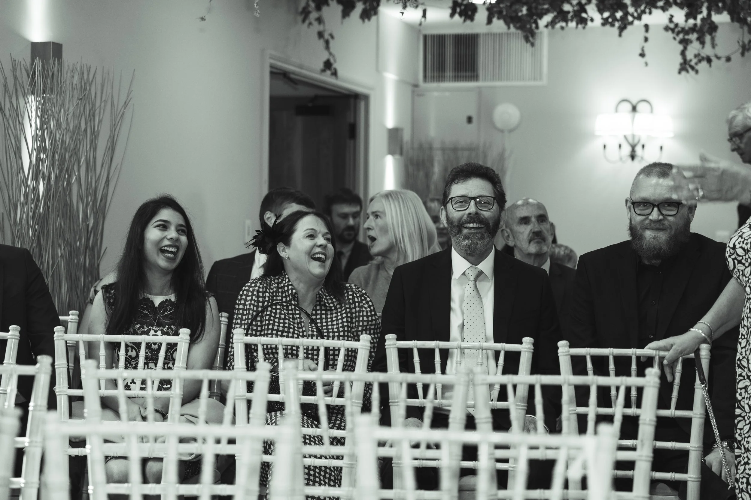 People sitting in chairs at an event, smiling and laughing, with a woman standing and handing out something to a bearded man in glasses.