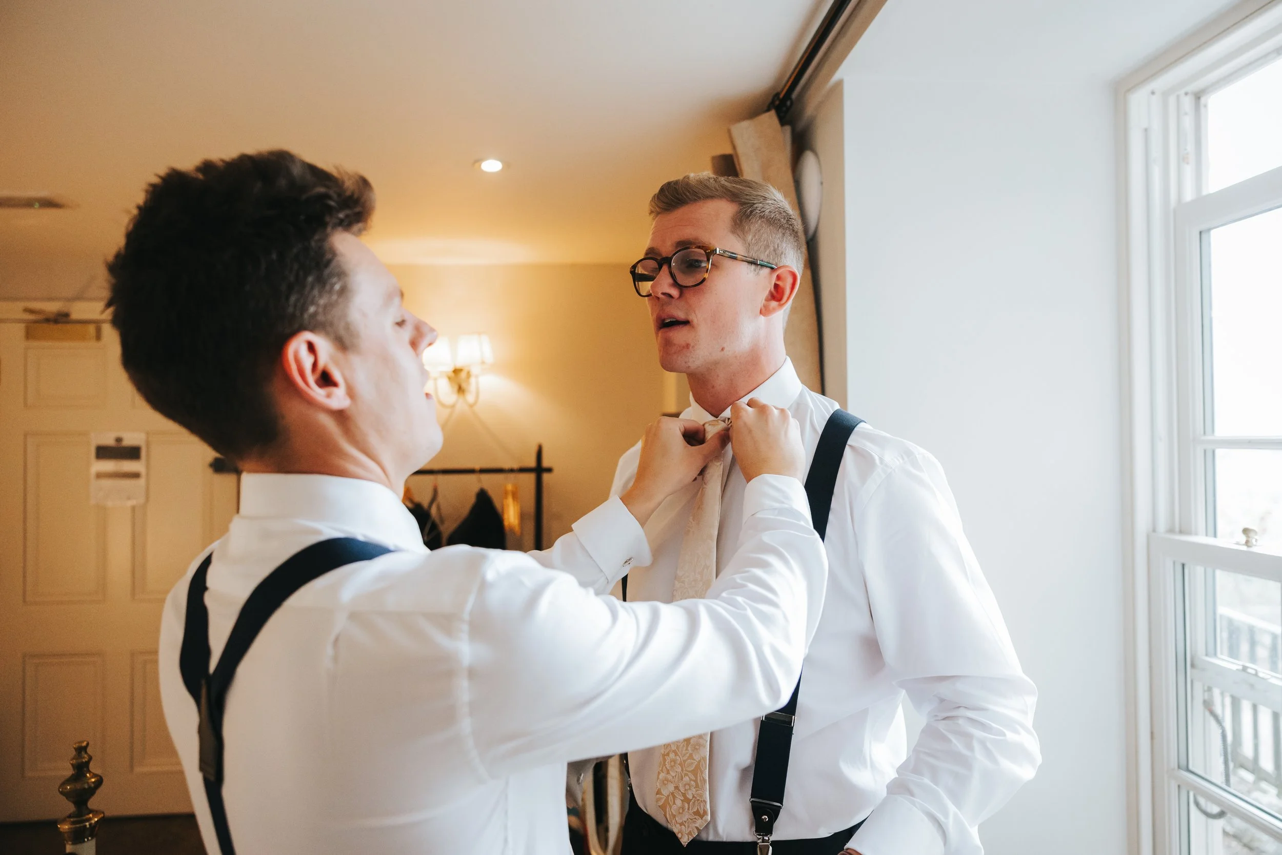 Two men in white shirts with suspenders, one helping the other adjust a tie near a window with natural light.