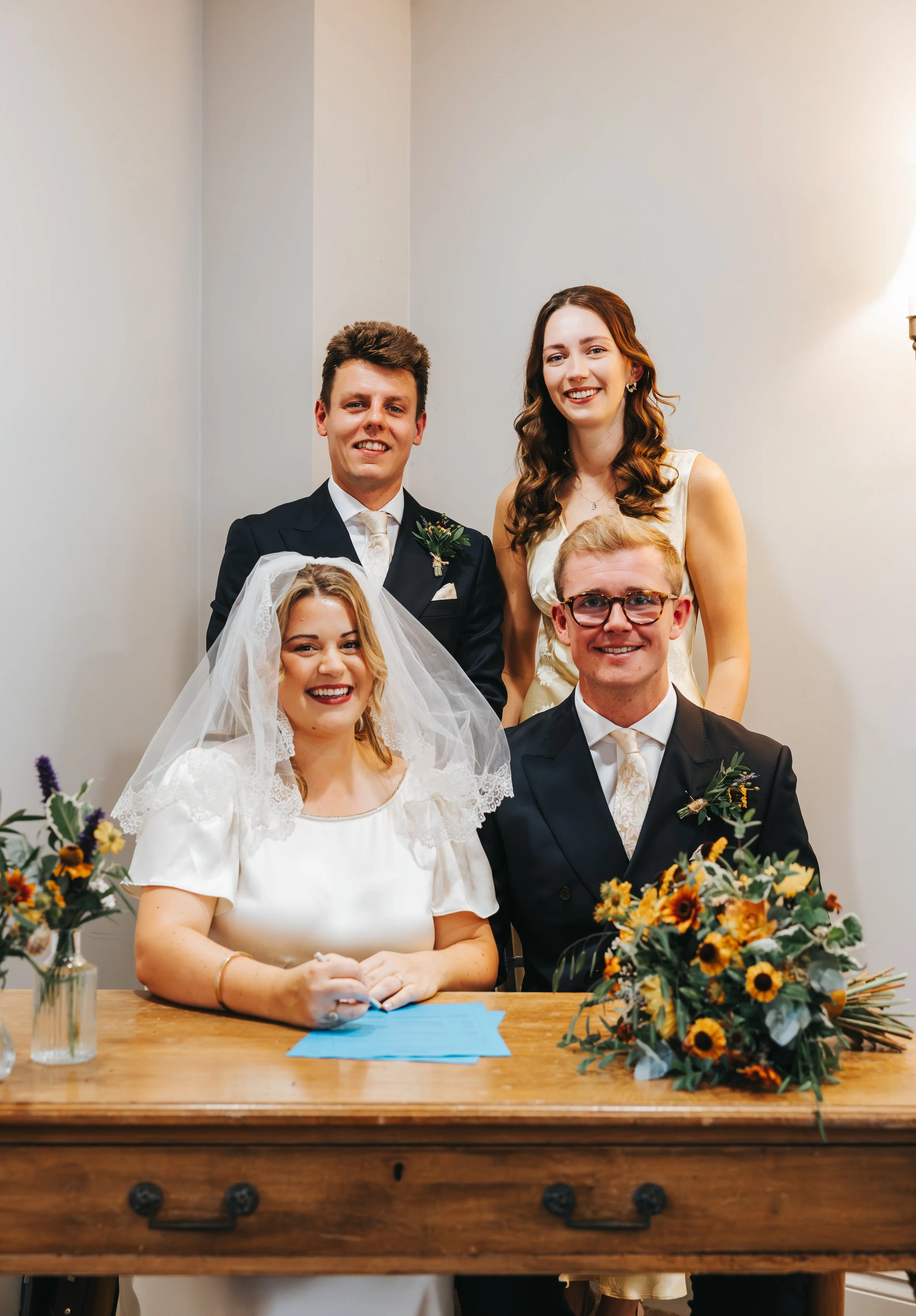 A wedding celebration with a bride and groom sitting at a table, surrounded by friends or family, with floral arrangements on the table.