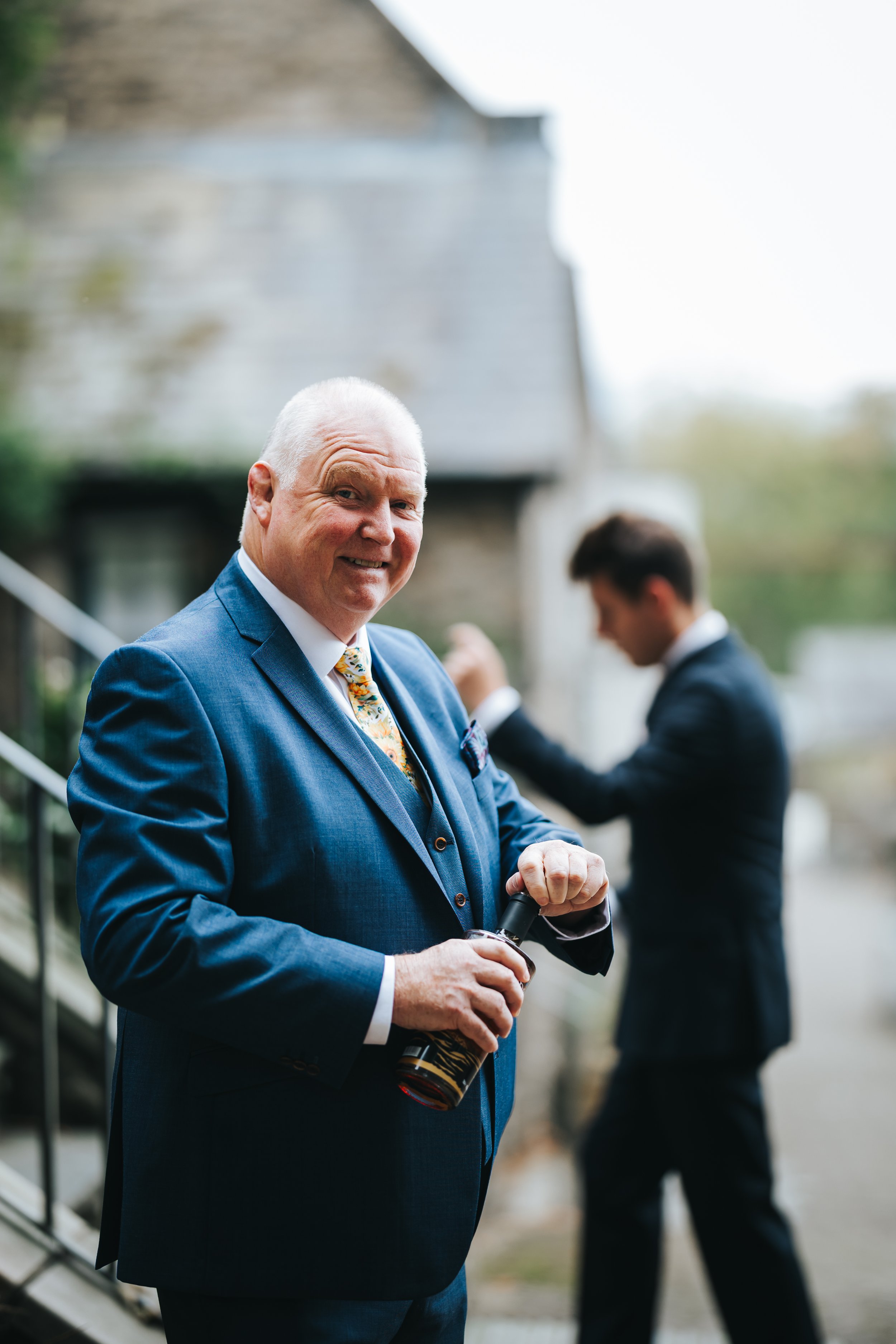 An elderly man in a blue suit holding a bottle of beer, smiling at the camera. In the background, a younger man in a dark suit is adjusting his tie.