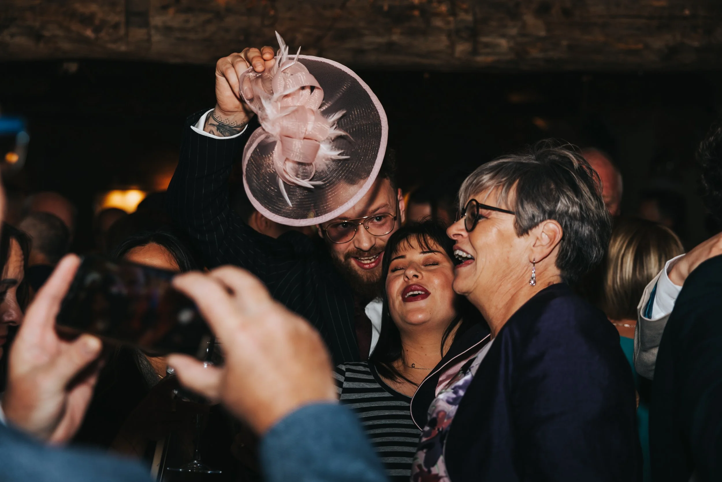 Group of people at a celebration, smiling, with one woman wearing large glasses, another woman in a floral dress, and a man holding a pink fascinator hat over his head.