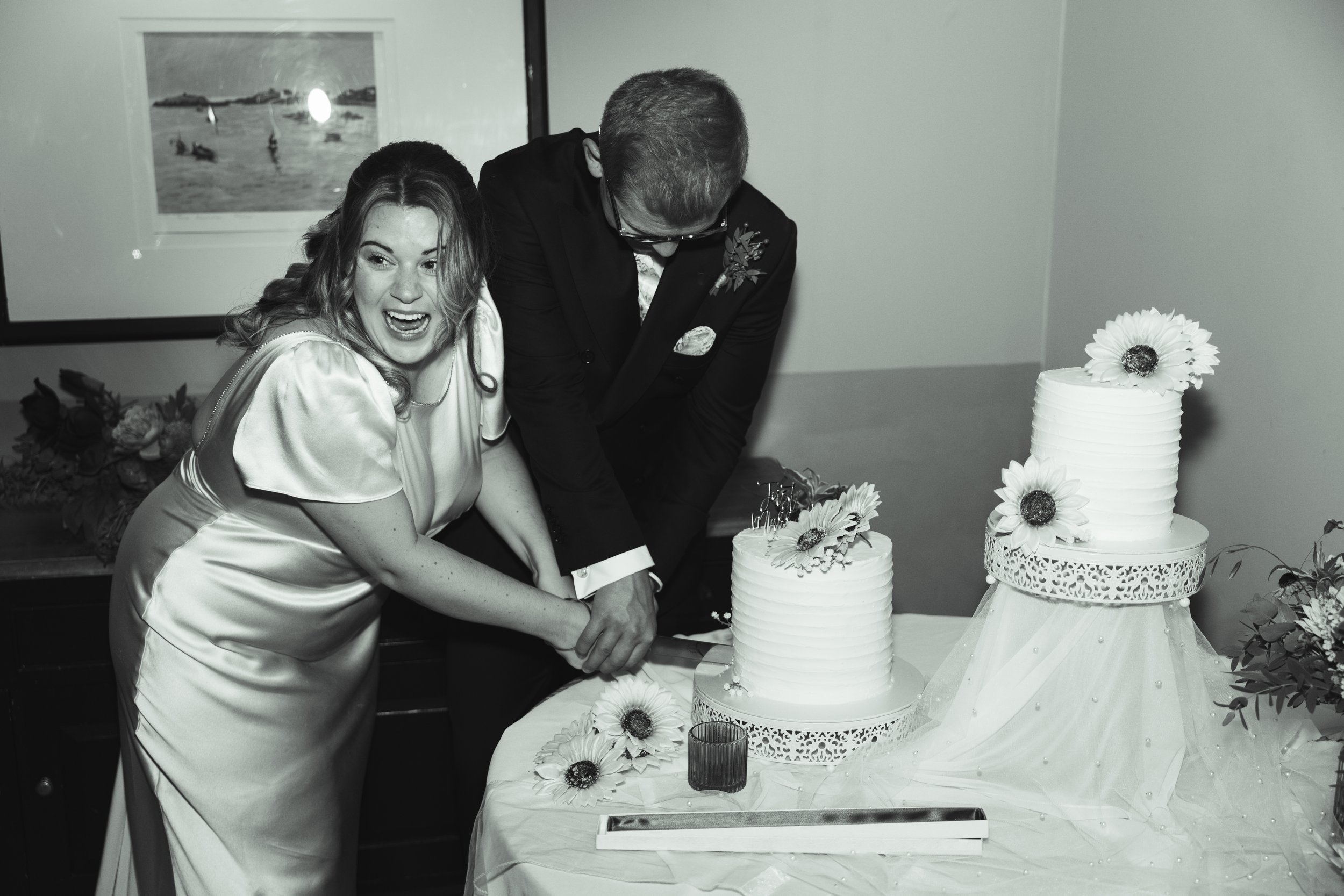 A couple is cutting a wedding cake together, the woman appears joyful and smiling, and both are dressed in formal wedding attire, with wedding decorations and flowers nearby.