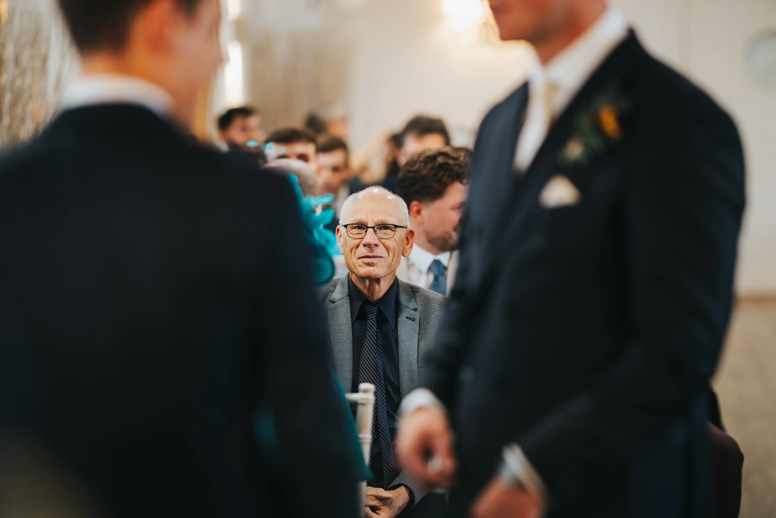 A man seated, watching a wedding ceremony between two men in suits and ties, with multiple guests in the background.