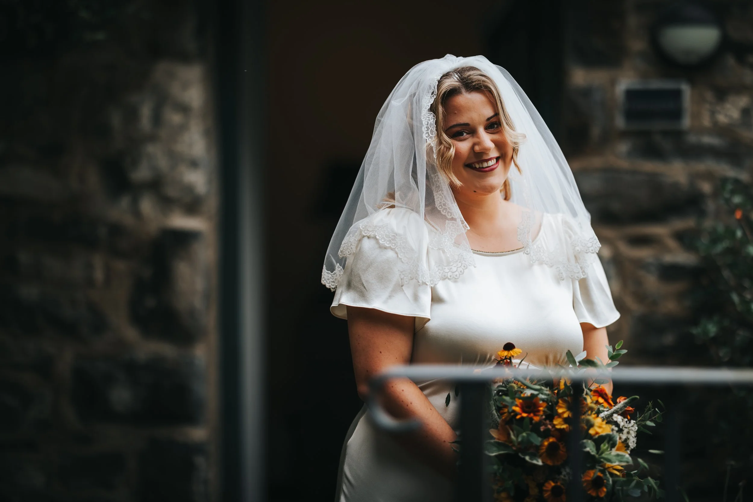 A smiling bride in a white wedding dress with lace accents and a veil, holding a bouquet of yellow and orange flowers, standing outdoors with a stone wall in the background.