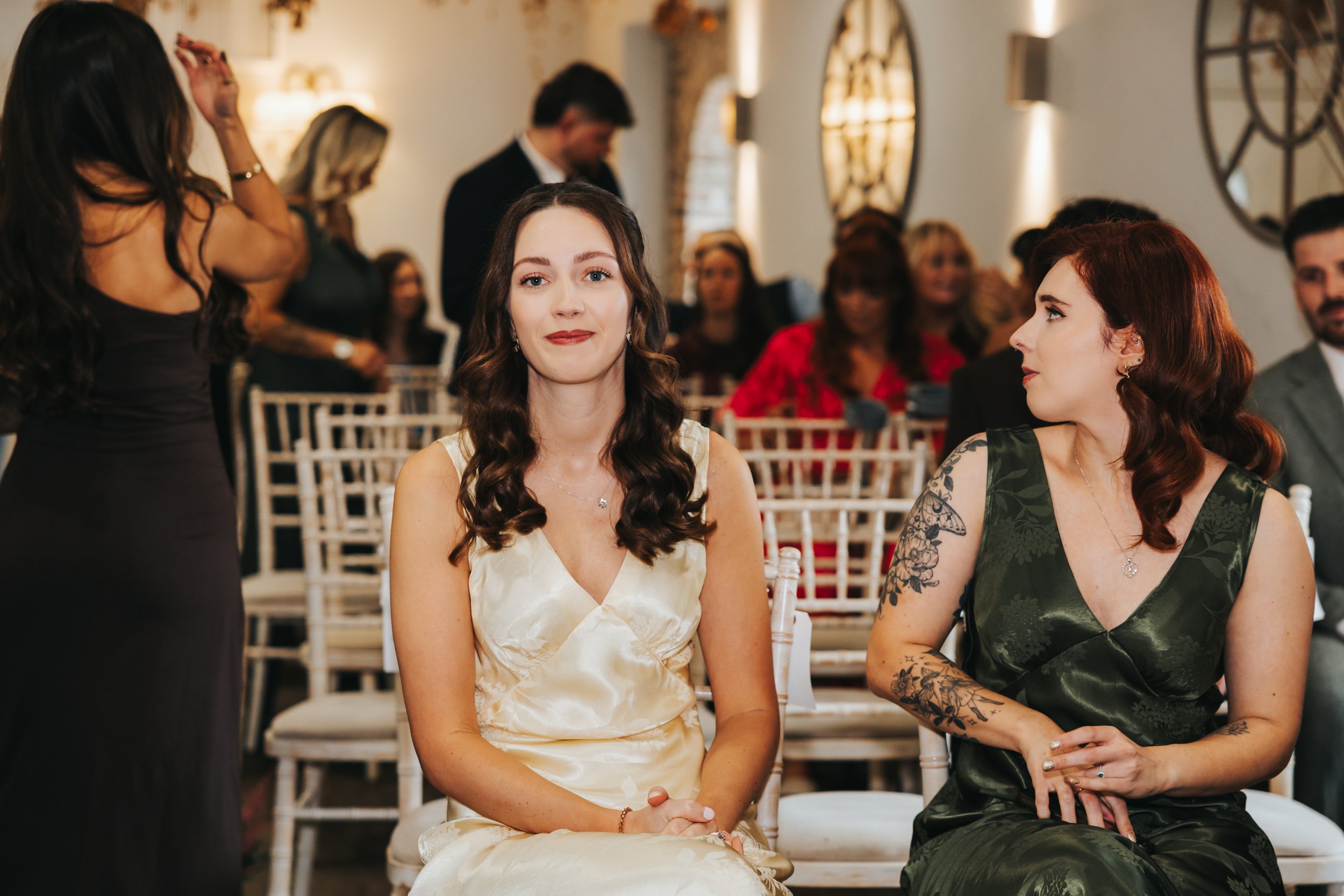 A woman with dark brown hair styled in loose curls, wearing a cream-colored satin dress, sits in a seated position at a formal event. Next to her, a woman with red hair and visible tattoos on her arm, wearing a dark green satin dress, also seated. In