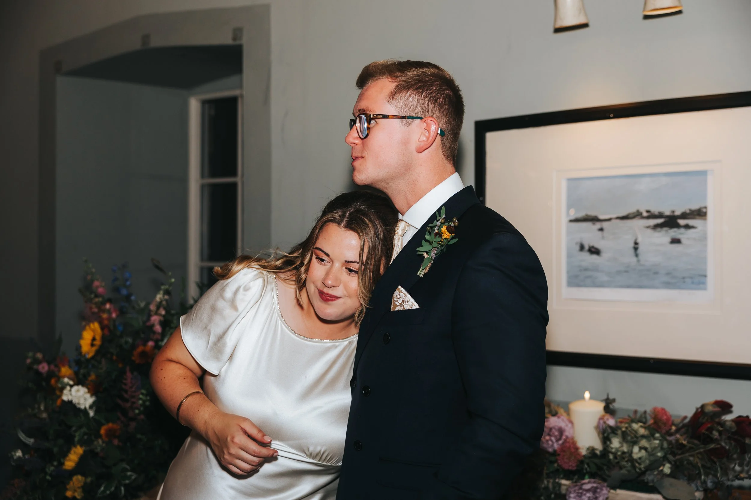 Two people at a wedding or formal event, a woman with shoulder-length brown hair leaning her head on a man in a dark suit with glasses, in front of a floral arrangement and artwork on the wall.