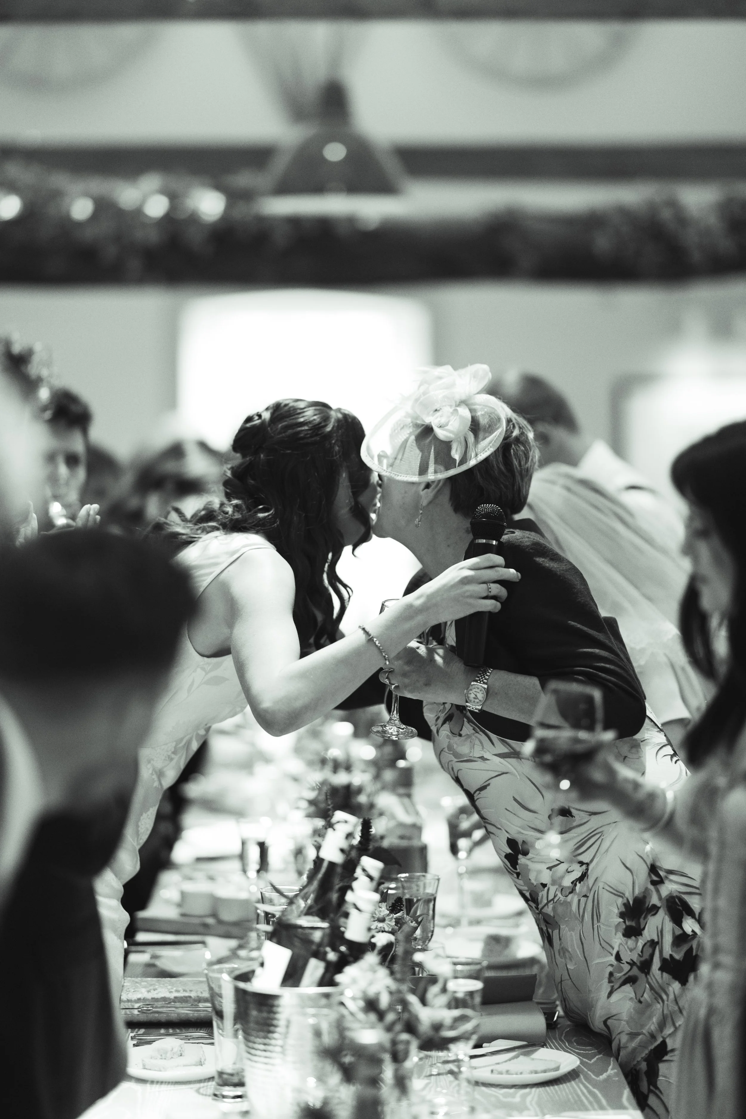 A black-and-white photo of people at a wedding reception, where a woman in a dress is kissing a woman with a fascinator hat, holding a microphone, at a decorated banquet table.