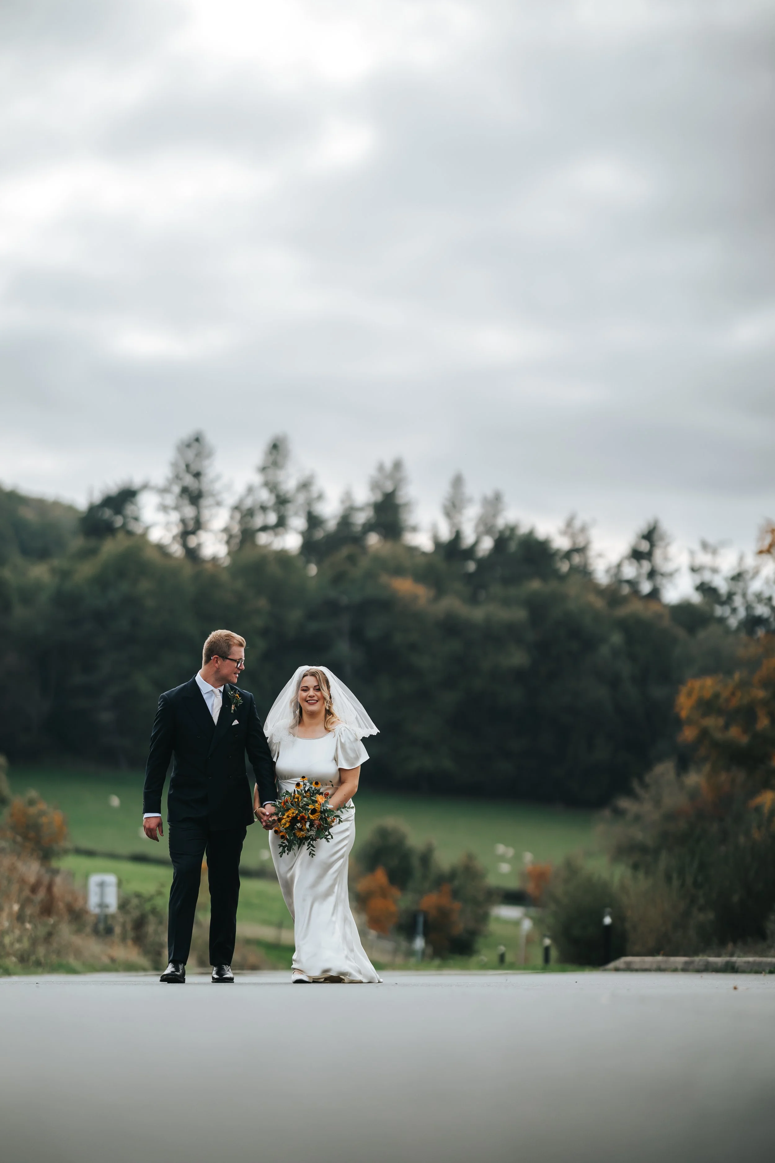 Bride and groom walking outdoors on a cloudy day, holding hands, with the bride holding a bouquet of flowers.