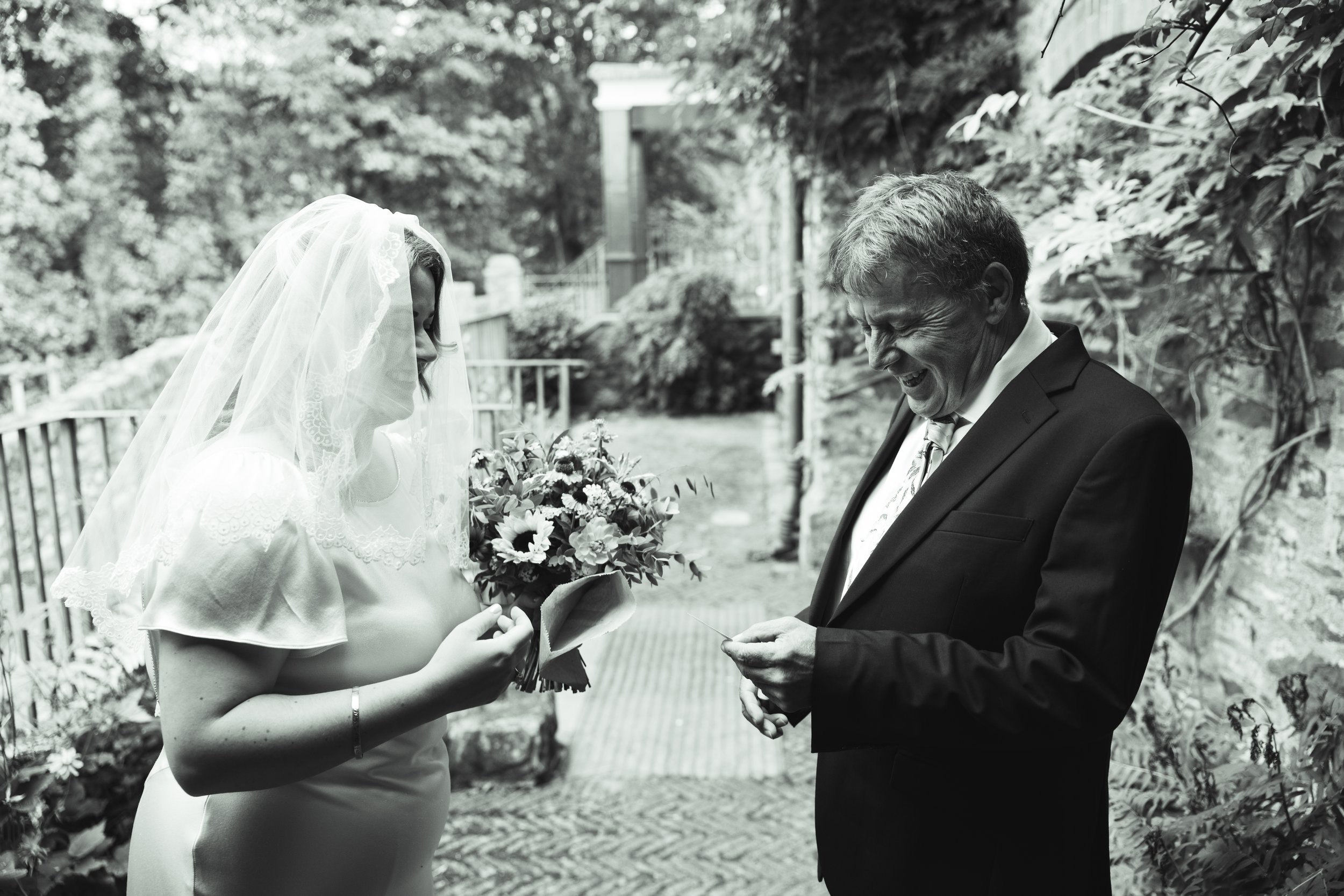 A bride and an older man, possibly her father, sharing a joyful moment outdoors. The bride is holding a bouquet of flowers, wearing a wedding dress and a veil, while the man is in a suit. Both are smiling and engaged in a happy interaction surrounded