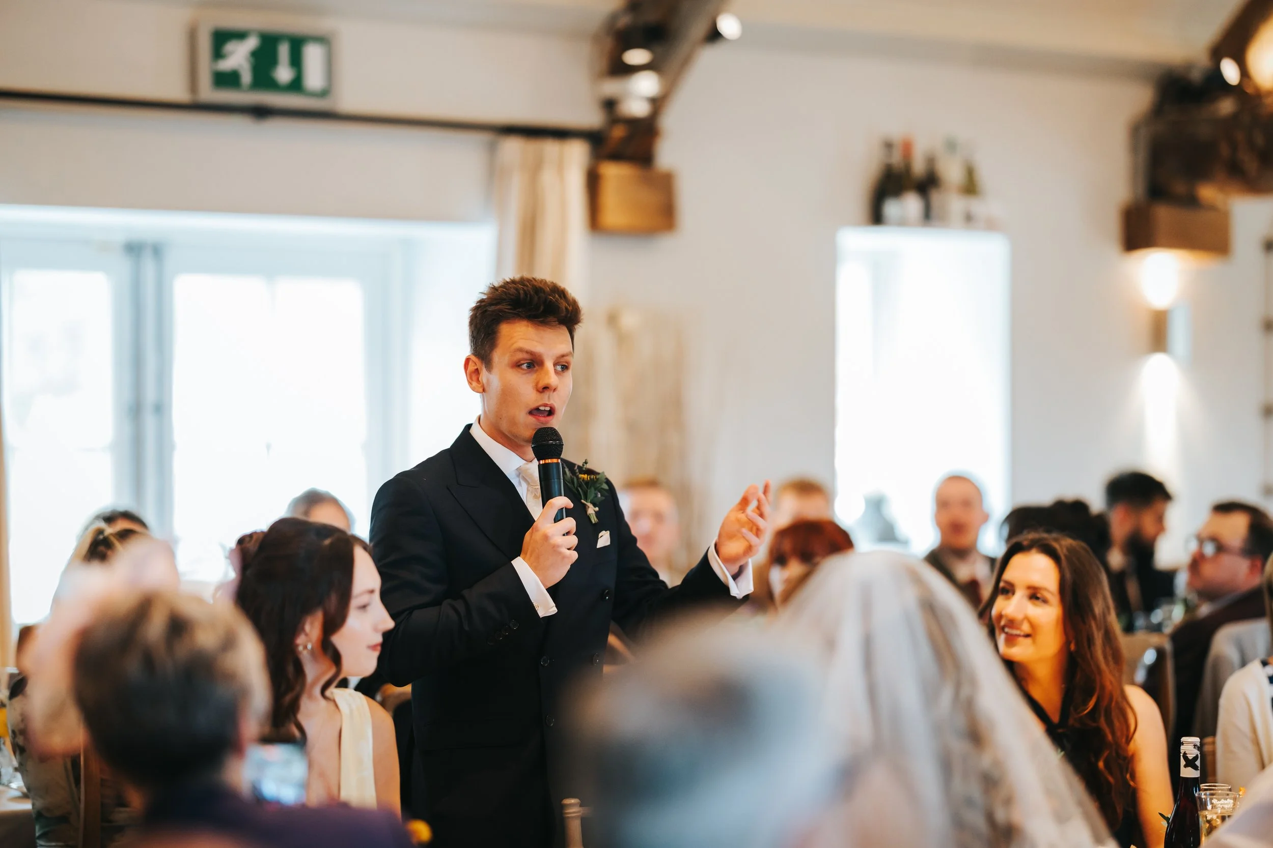 A young man in a tuxedo giving a speech with a microphone at a wedding reception, surrounded by seated guests in a rustic decorated venue with large windows.