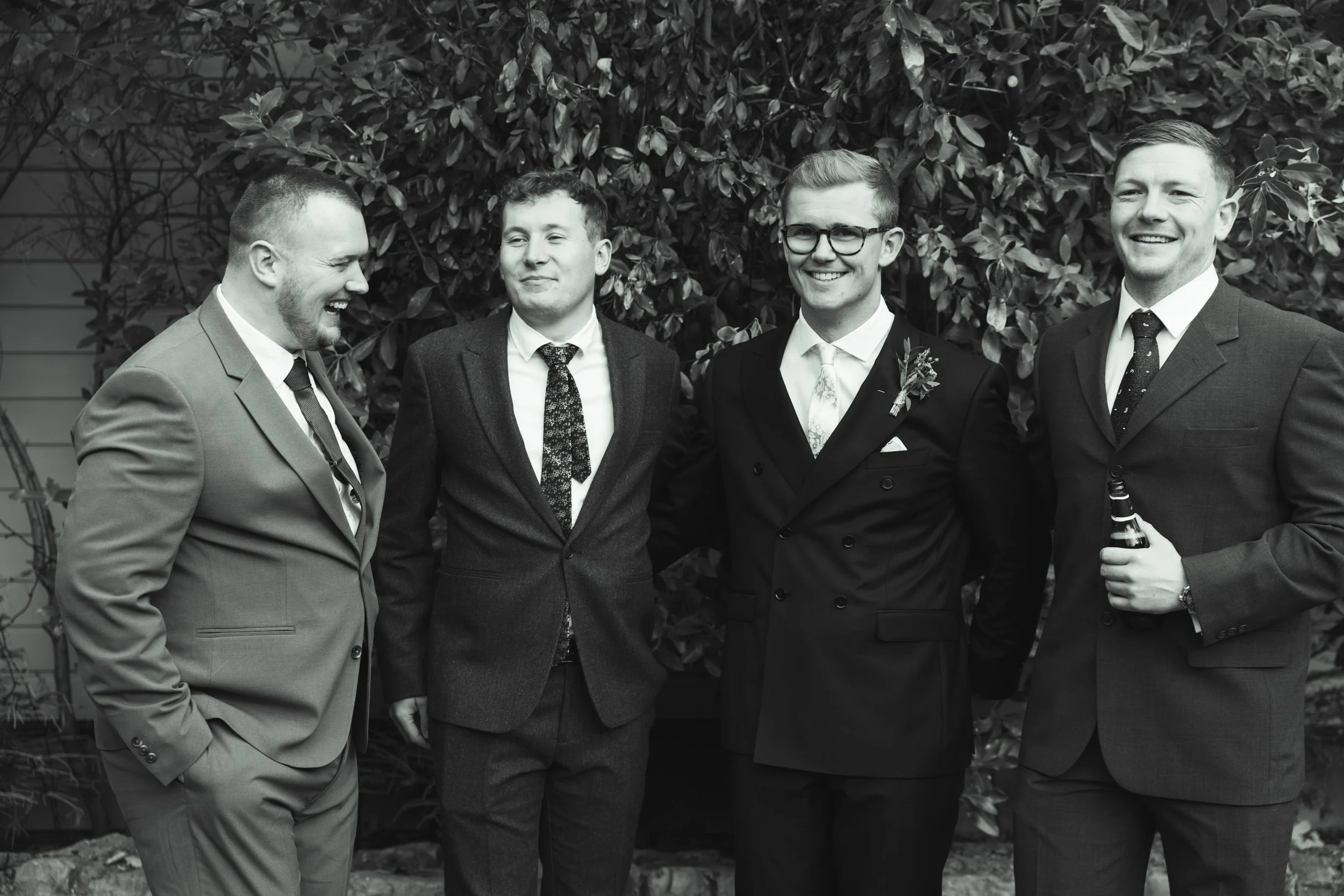 Four men in suits standing together outdoors, smiling and laughing, with a leafy background, celebrating a wedding or special occasion.