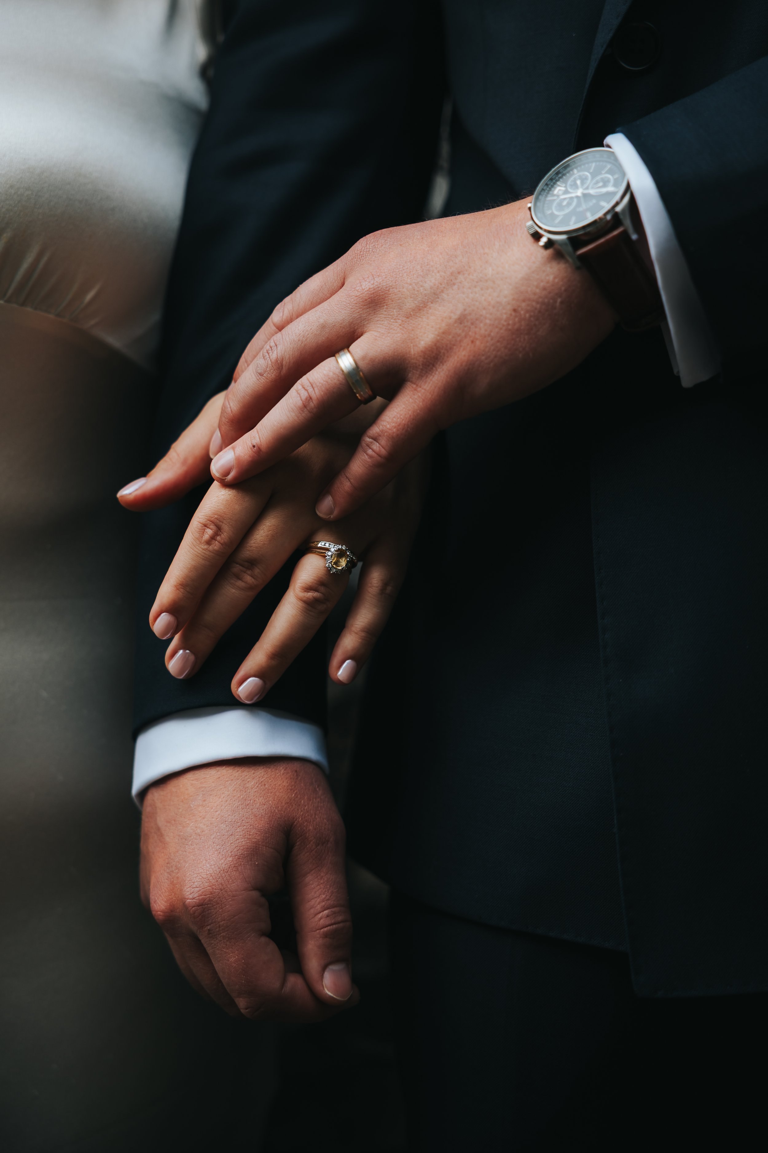 Close-up of a man and woman holding hands, both wearing wedding rings and the man wearing a watch, dressed in formal attire.