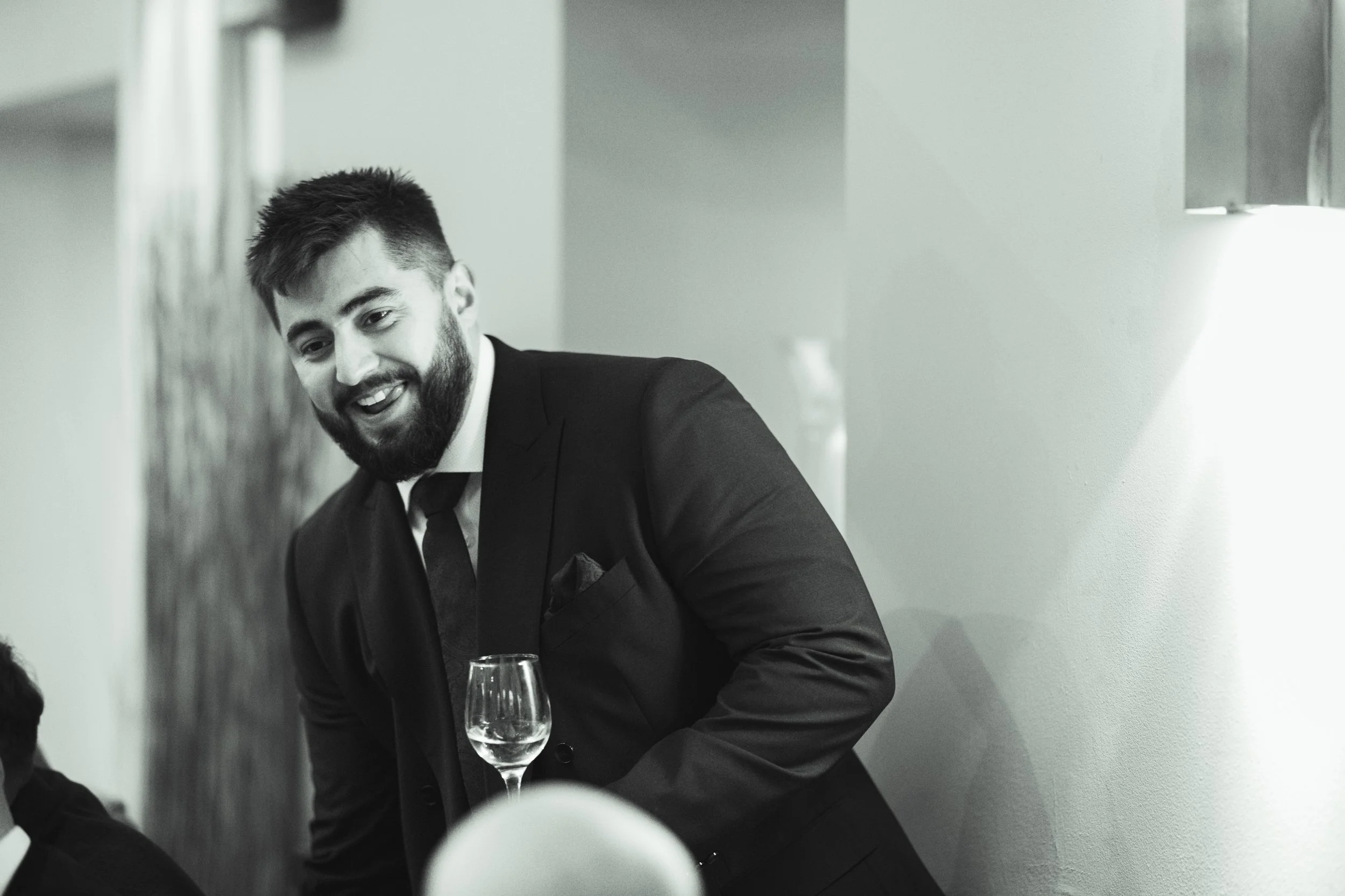 Man with a beard in a suit smiling and pouring wine at a social gathering.