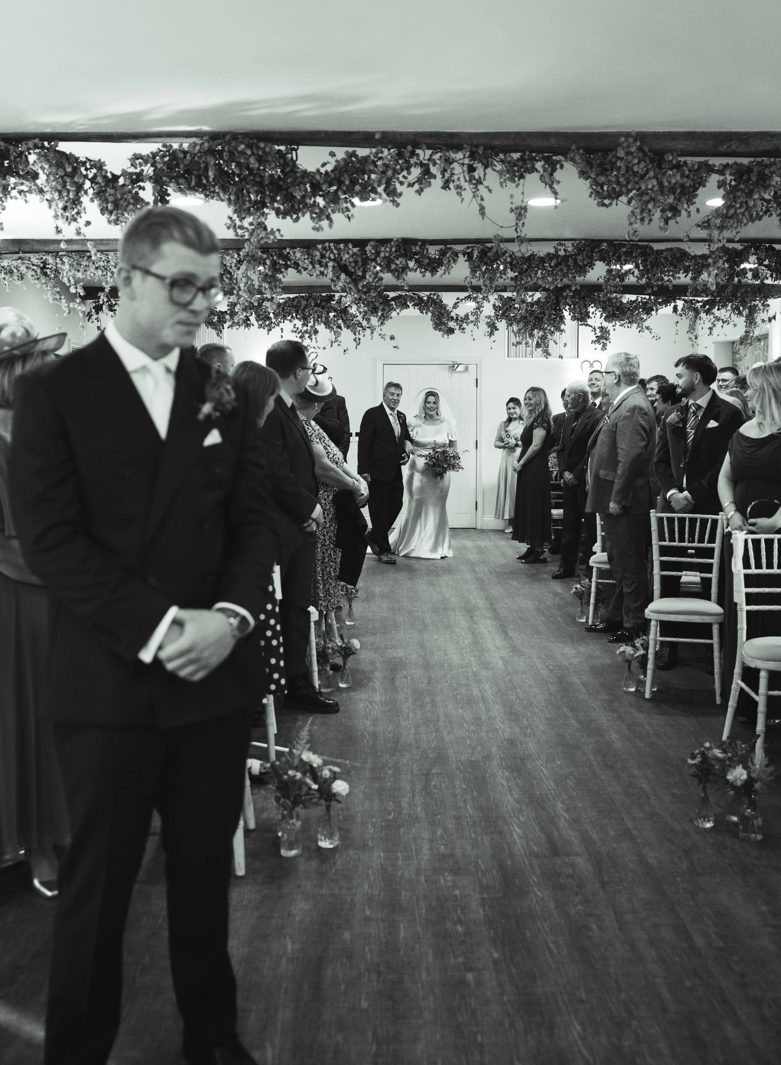 Black and white photo of a wedding ceremony with guests standing on either side of the aisle. A bride in a white dress holding flowers stands at the front, smiling, with an older man next to her. The ceiling is decorated with hanging flowers.