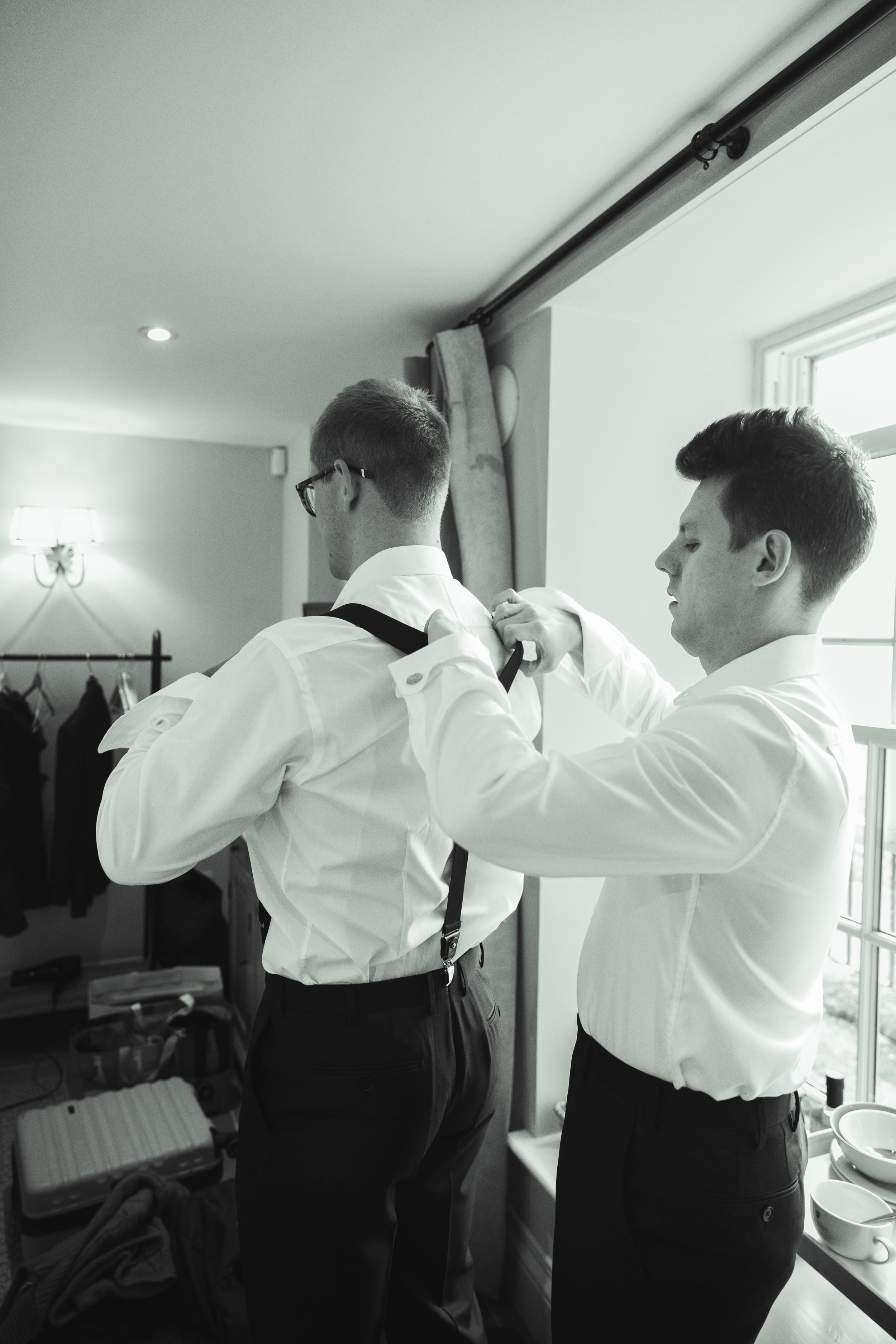 Two men in white shirts and black pants are helping each other, with one adjusting the other's suspenders in a well-lit room near a window.