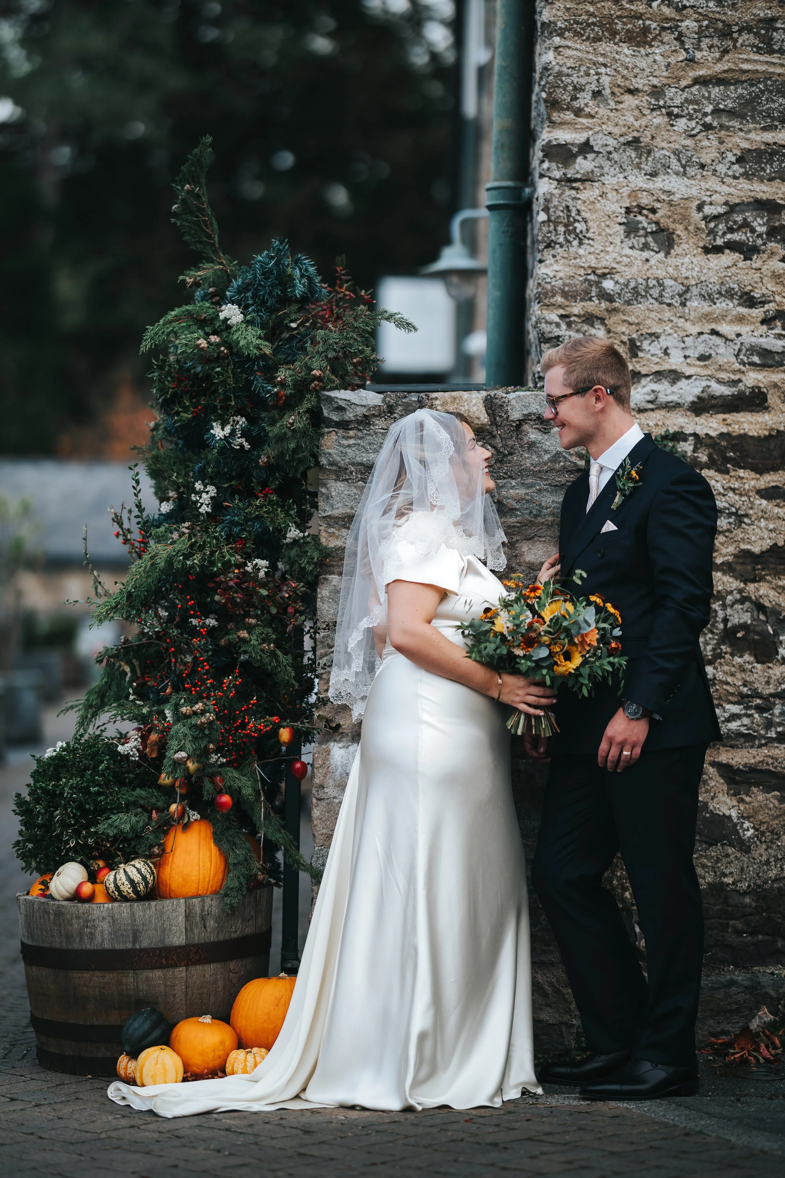 A bride and groom looking at each other and smiling during their wedding in front of a stone wall decorated with fall pumpkins and a small Christmas tree.