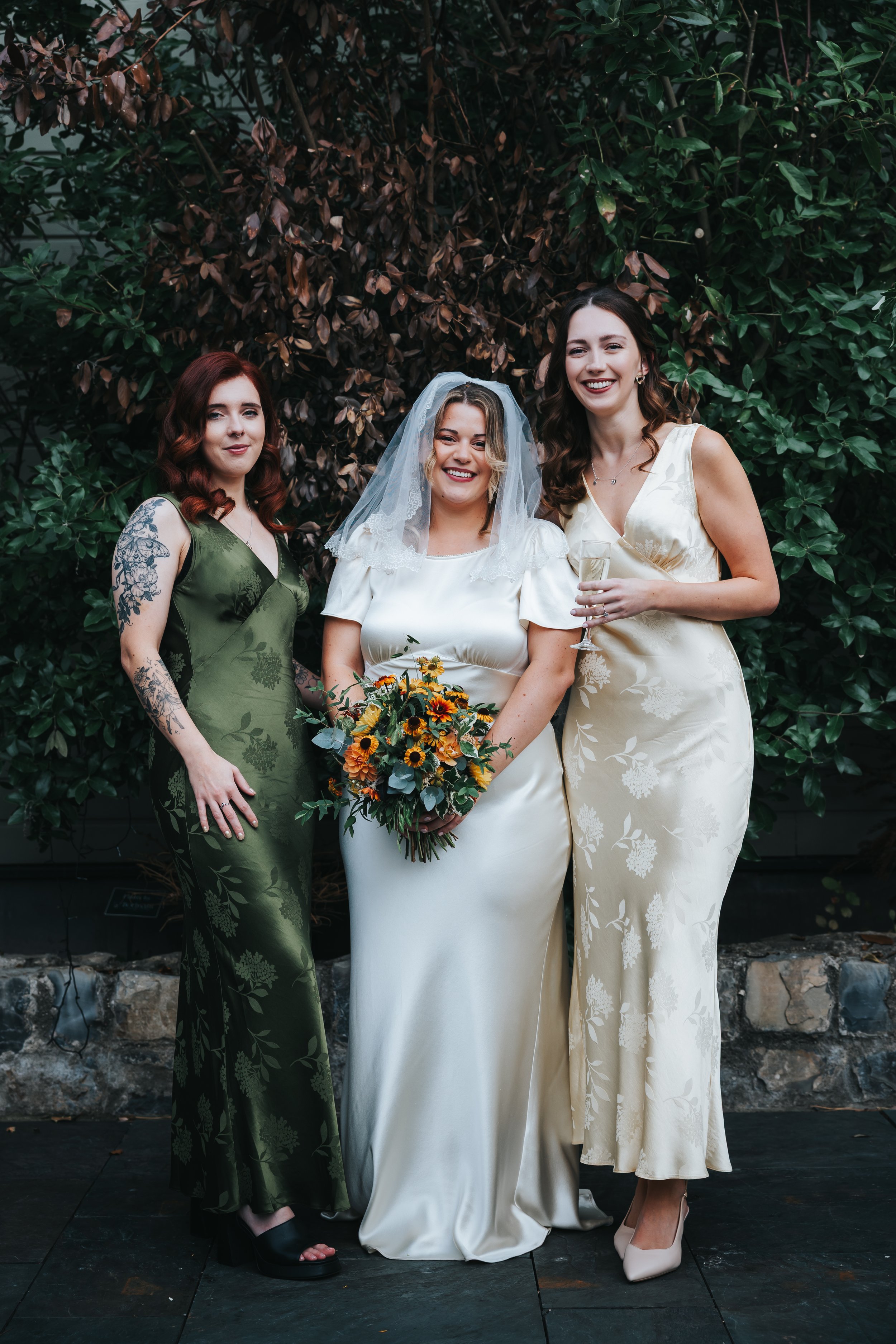Three women at a wedding, with the bride holding a bouquet of sunflowers, wearing a white wedding dress and veil, and two women in elegant dresses, one in green and the other in cream, standing outdoors in front of green and brown foliage.
