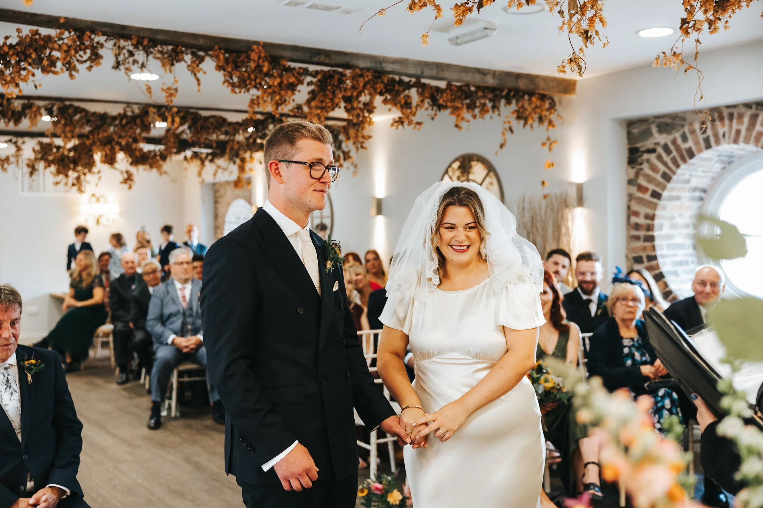 A bride and groom holding hands and smiling during their wedding ceremony indoors, with seated guests in the background and autumn-themed decorations hanging from the ceiling.