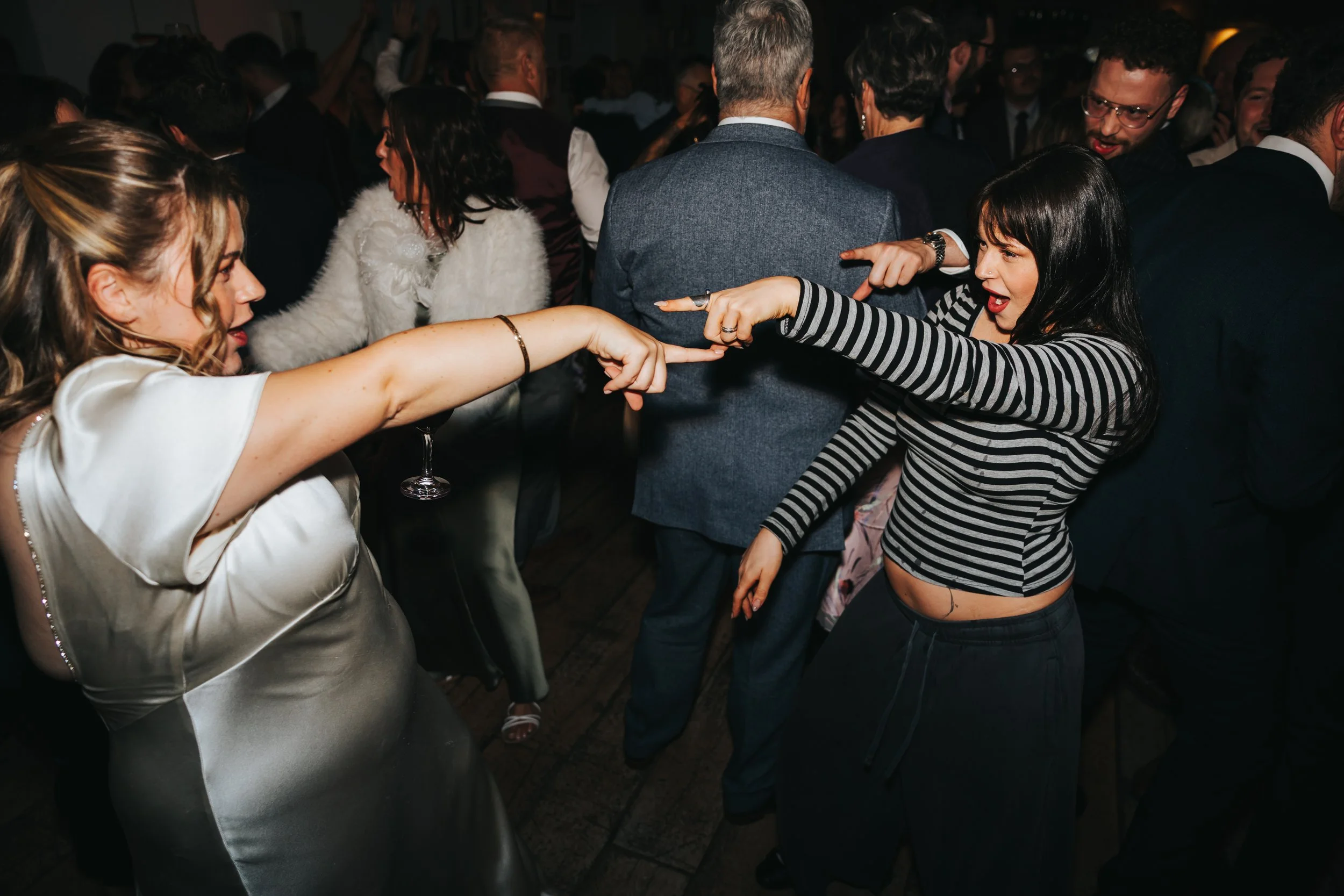 Two women dancing and pointing at each other at a lively party or event. One woman has blonde hair and is wearing a shiny, light-colored dress, while the other has dark hair and is dressed in a black and white striped top with black pants. The backgr