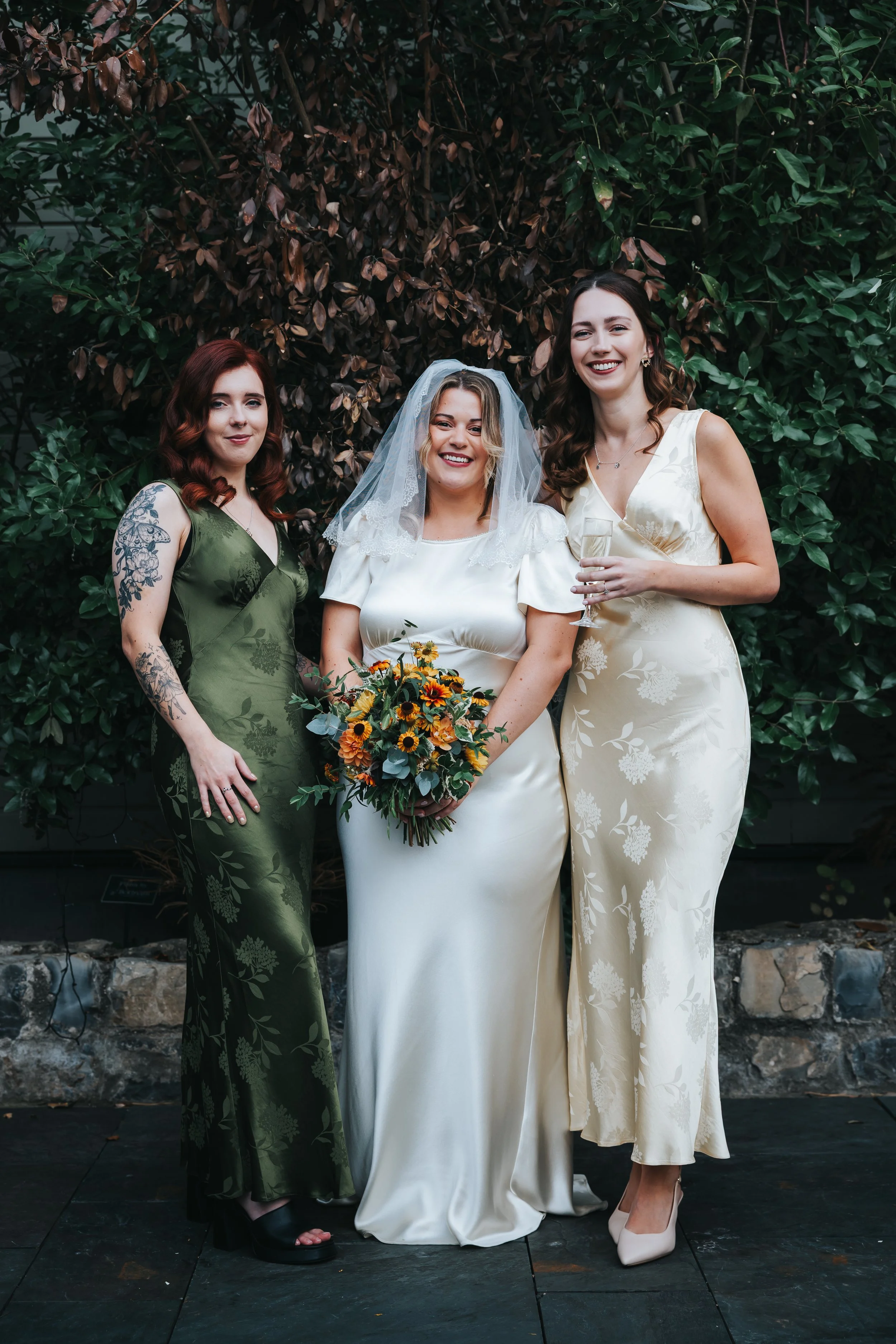 Three women at a wedding, one in a white wedding dress holding a bouquet, and two in evening gowns, standing outdoors in front of lush greenery.