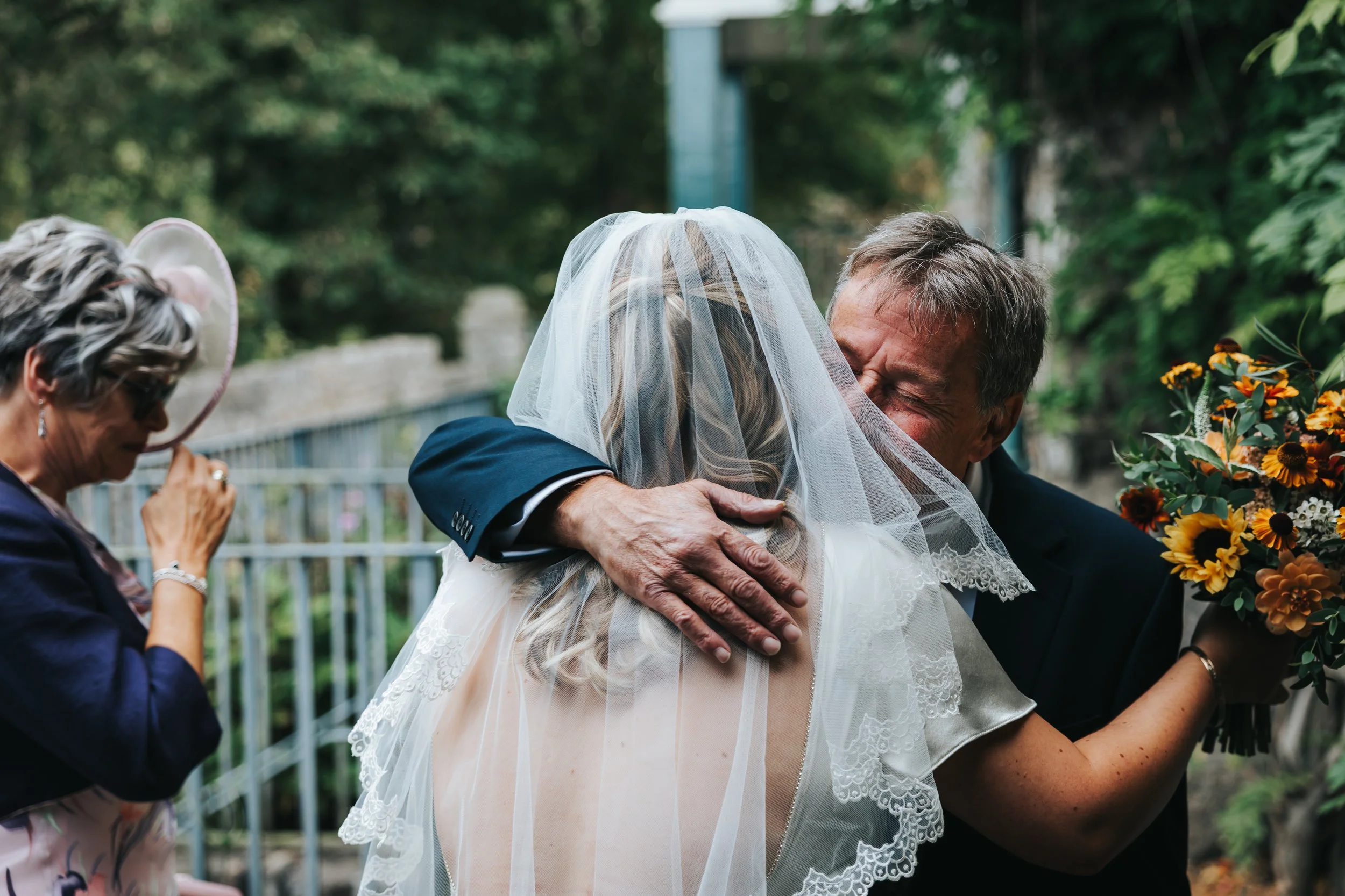 A bride hugging an older man, possibly her father, while a woman with a hat looks on in the background at an outdoor wedding.