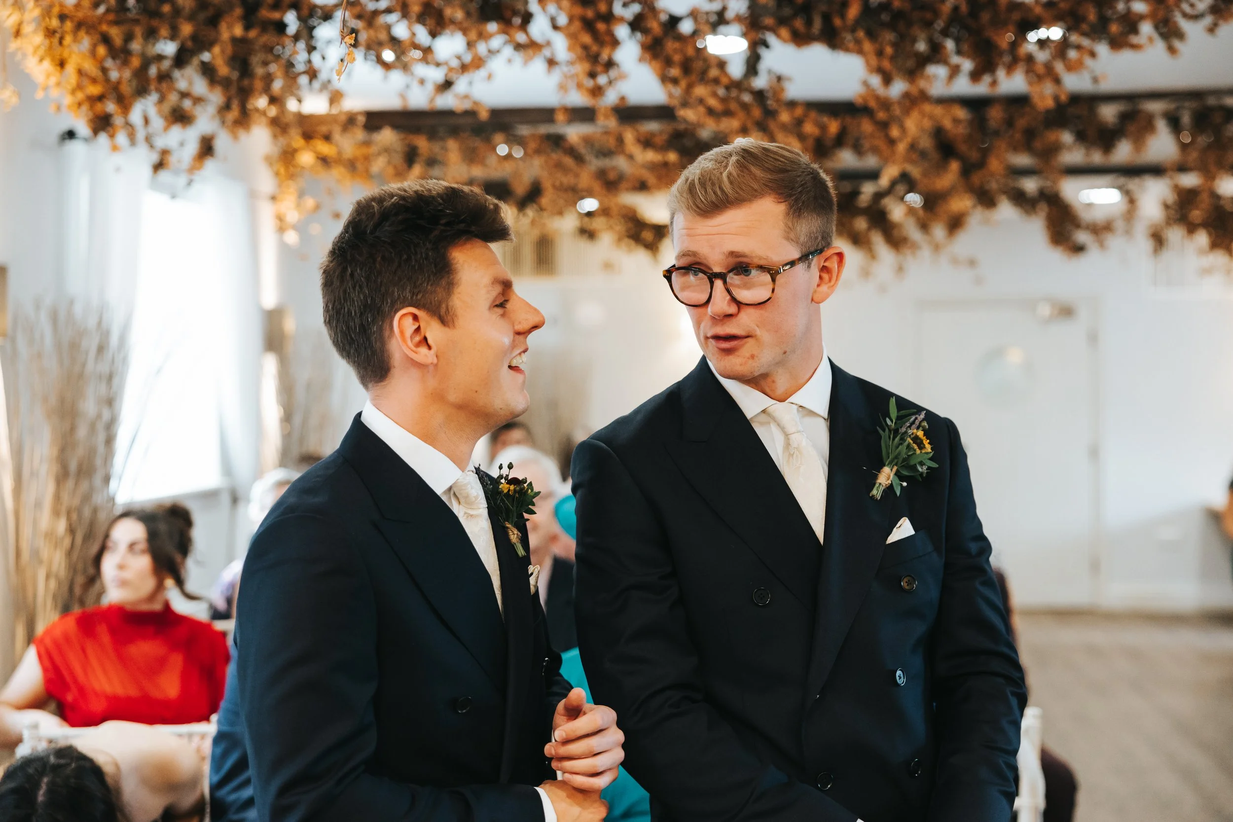 Two men in tuxedos talking at a wedding reception with autumn-themed decorations.