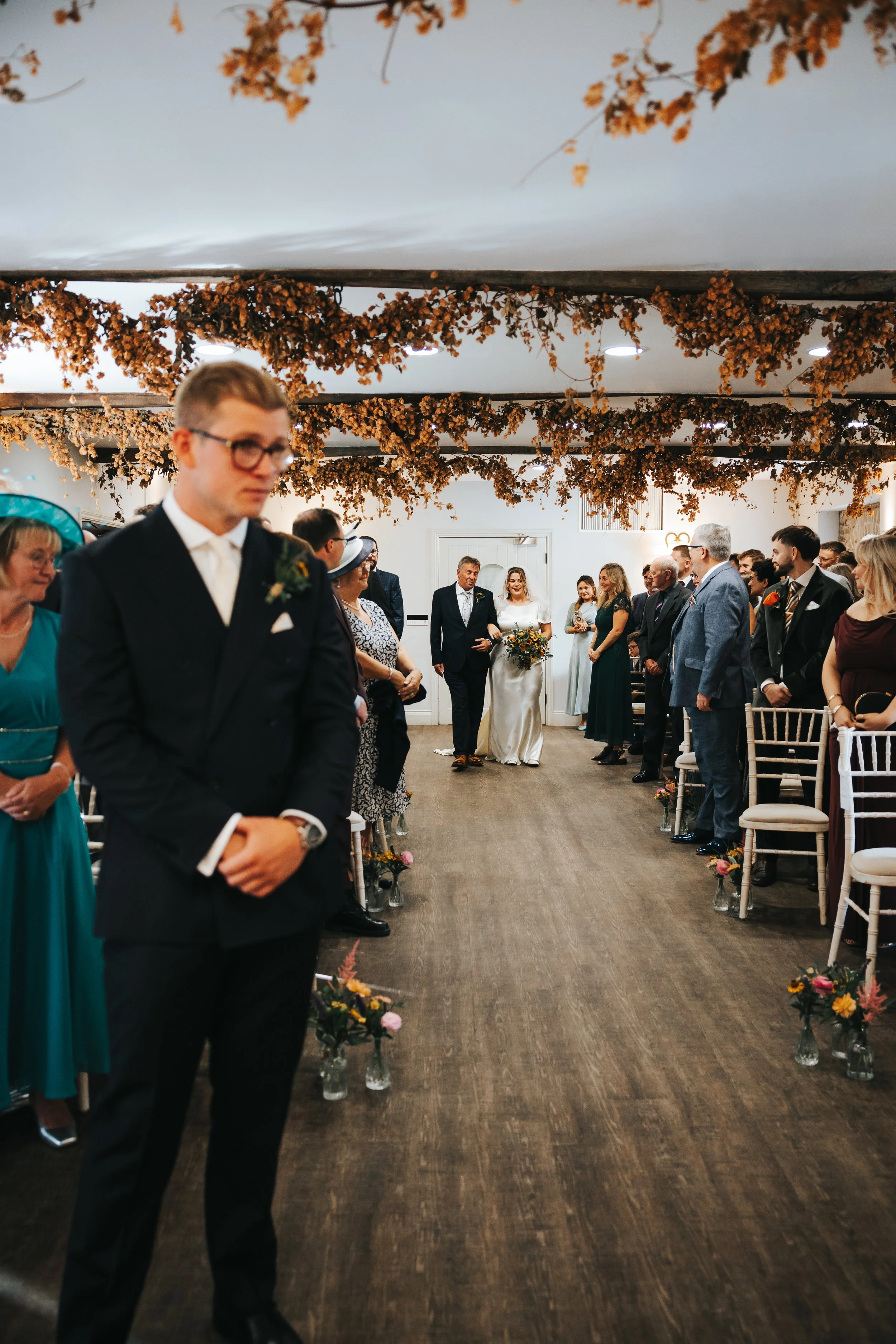 Wedding ceremony inside a decorated venue with hanging flower arrangements on the ceiling, guests seated on either side of an aisle, and a bride walking down the aisle with her father.