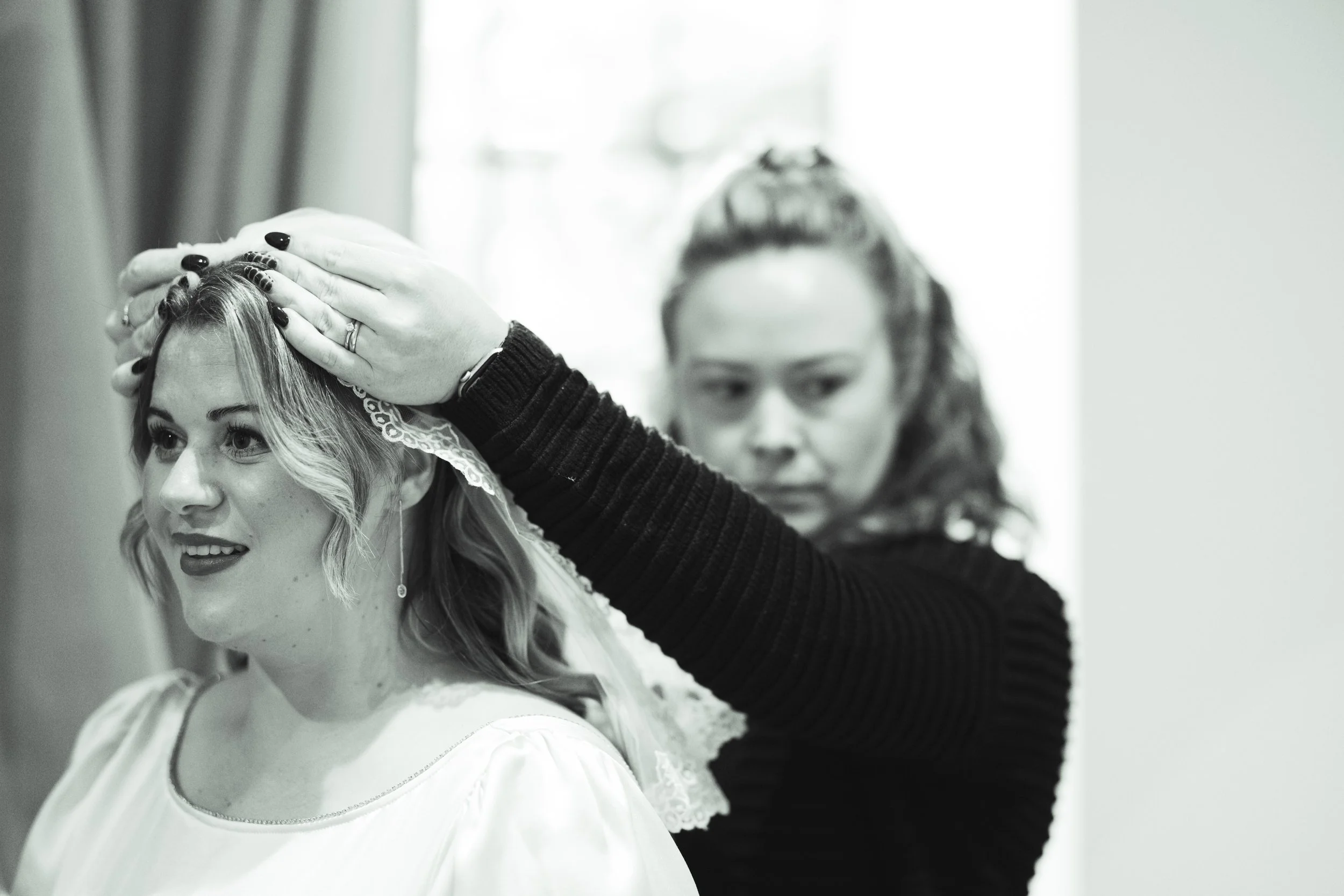 A woman with wavy hair is having a veil arranged on her head by another woman with curly hair, both focused on the task.