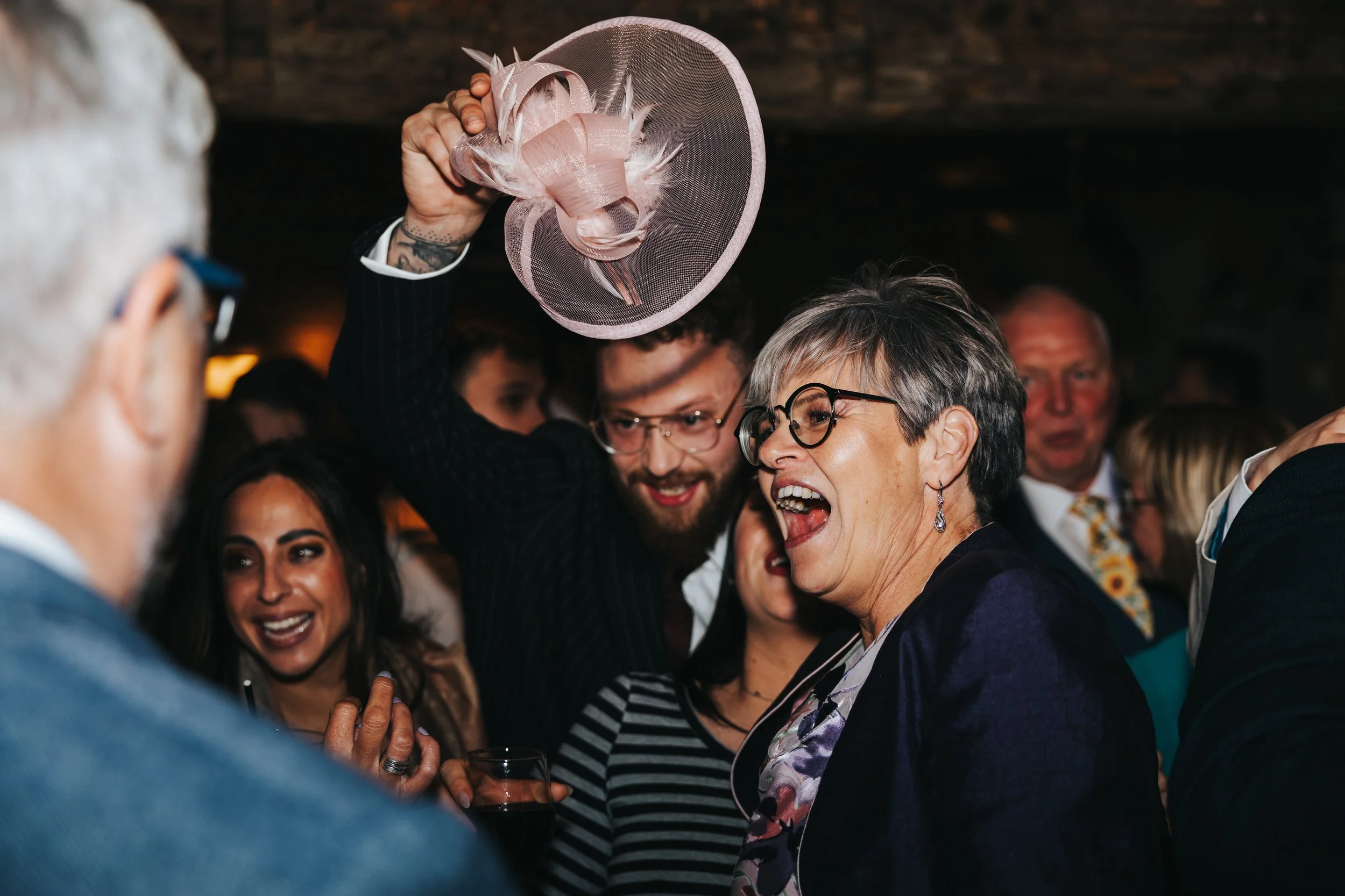 Group of people celebrating together at an indoor party, woman holding a pink hat with feathers, smiling and laughing