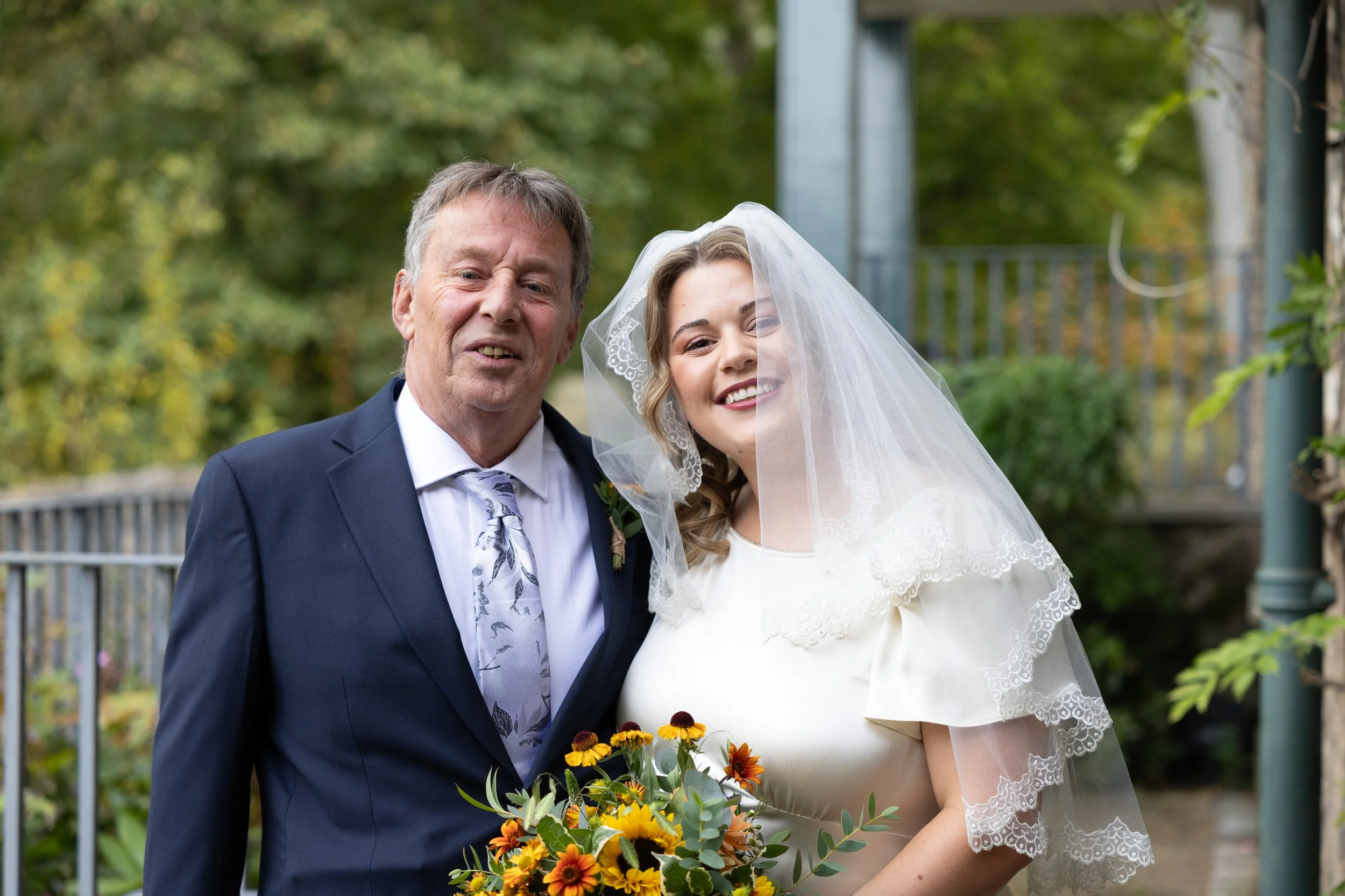 A bride in a white dress and veil holding a bouquet of yellow and orange flowers, standing next to an older man in a dark suit, both smiling outdoors with green trees in the background.