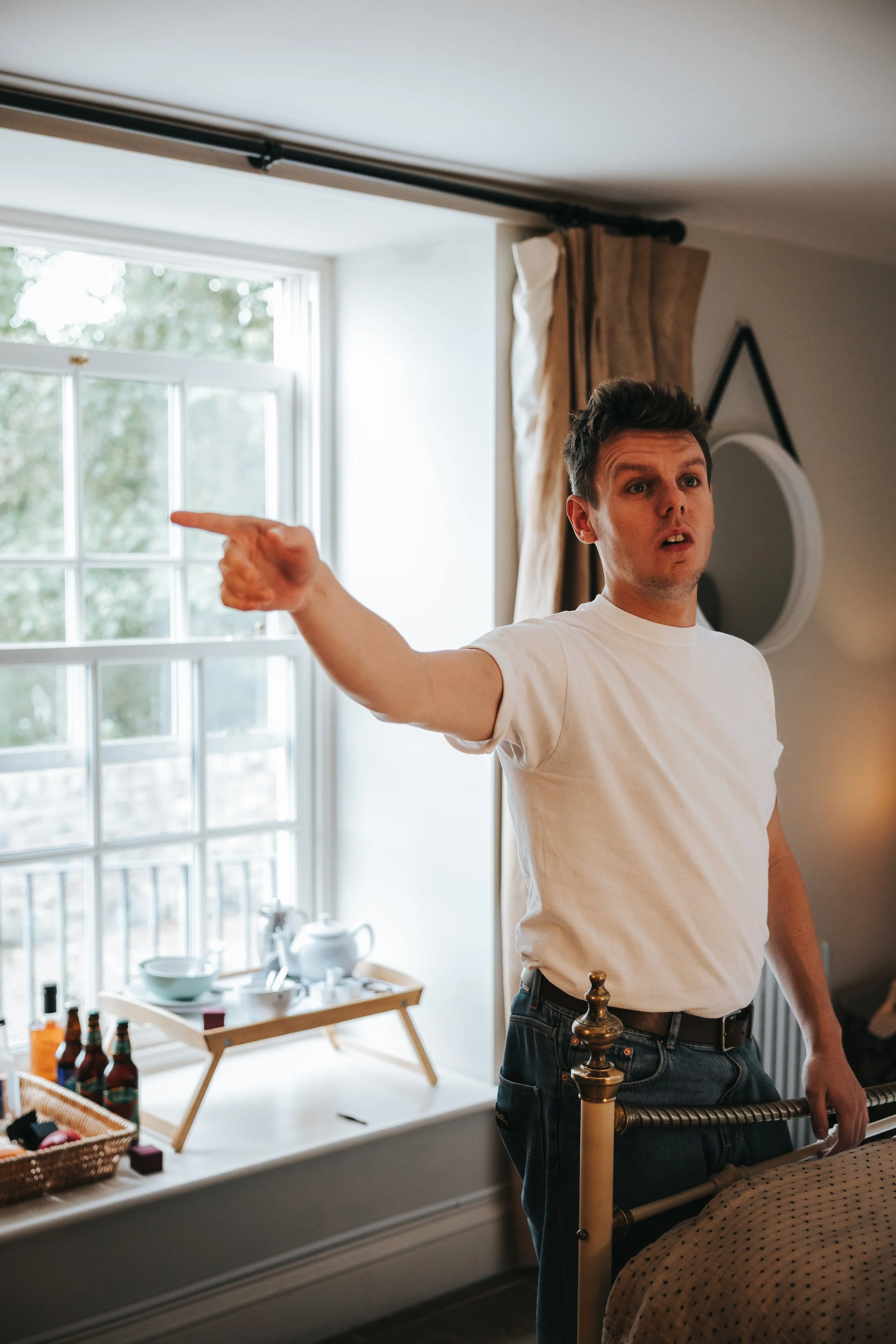 A man with short, dark hair is pointing to his left with a surprised or confused expression. He is wearing a white T-shirt and dark jeans, standing next to a bed in a well-lit room with a large window, brown curtains, and decorative items like bottle