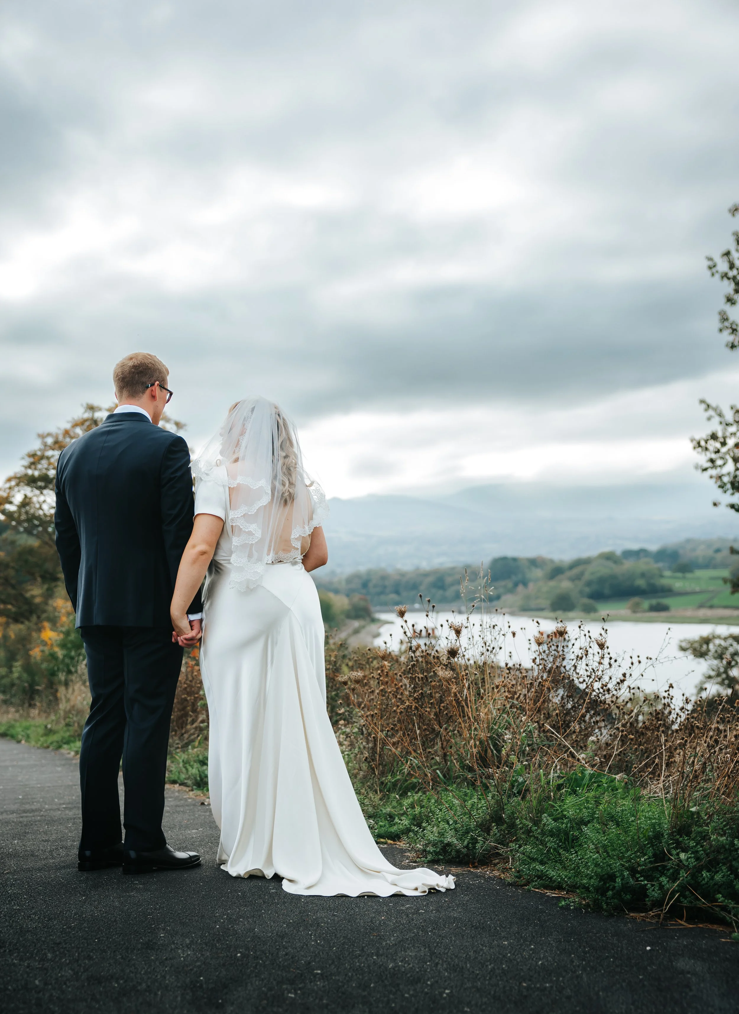 A bride and groom holding hands and standing outdoors on a cloudy day, overlooking a river and hills.