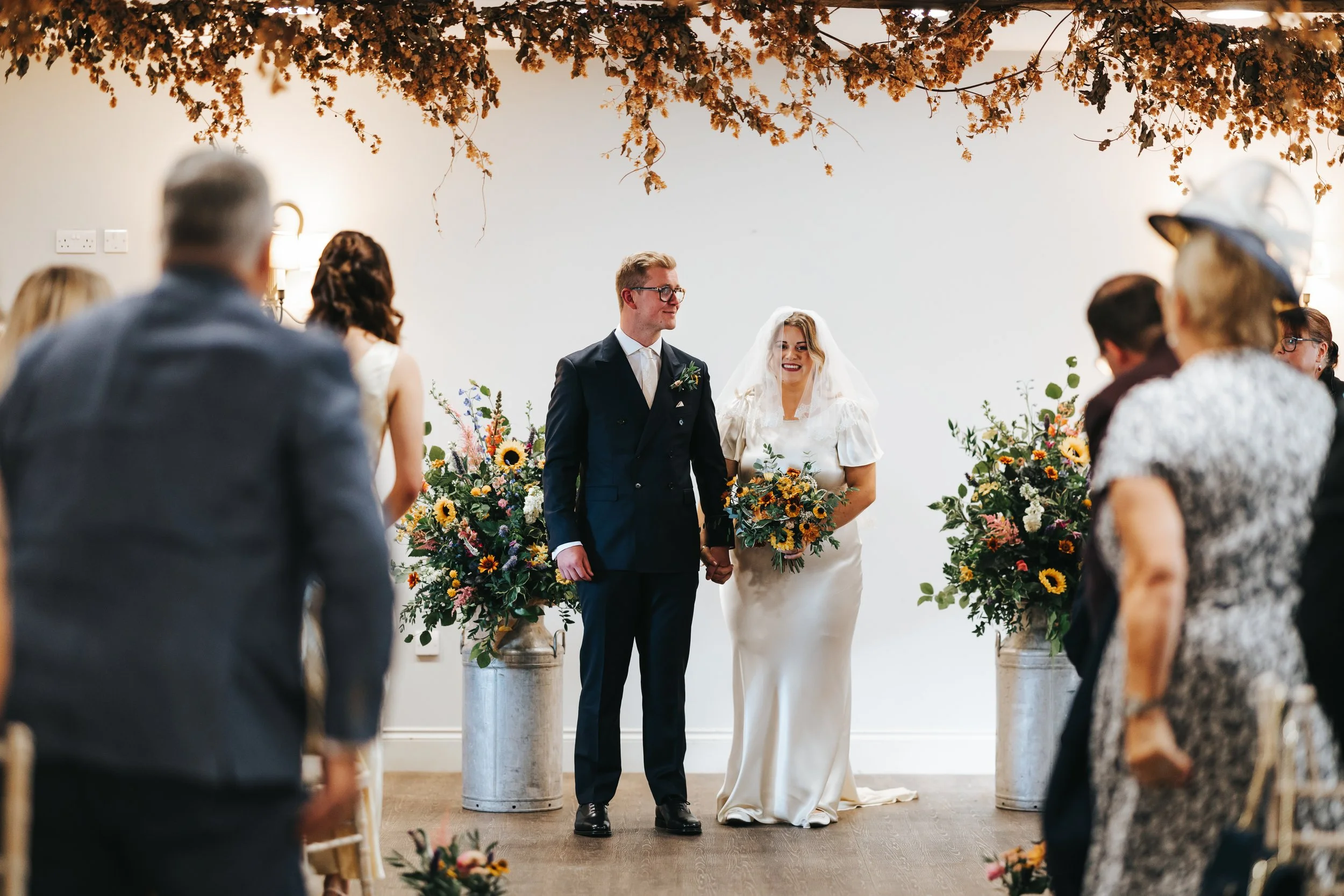 A bride and groom stand hand in hand during their wedding ceremony, surrounded by guests. The bride is wearing a white wedding dress with a veil, holding a bouquet of flowers, and the groom is dressed in a dark suit with glasses. There are large flor