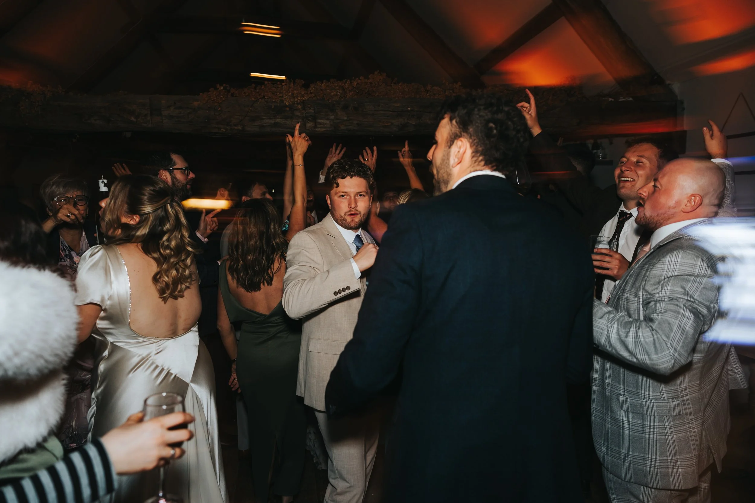 People dancing and celebrating at a party or wedding reception in a dimly lit room with warm lighting.