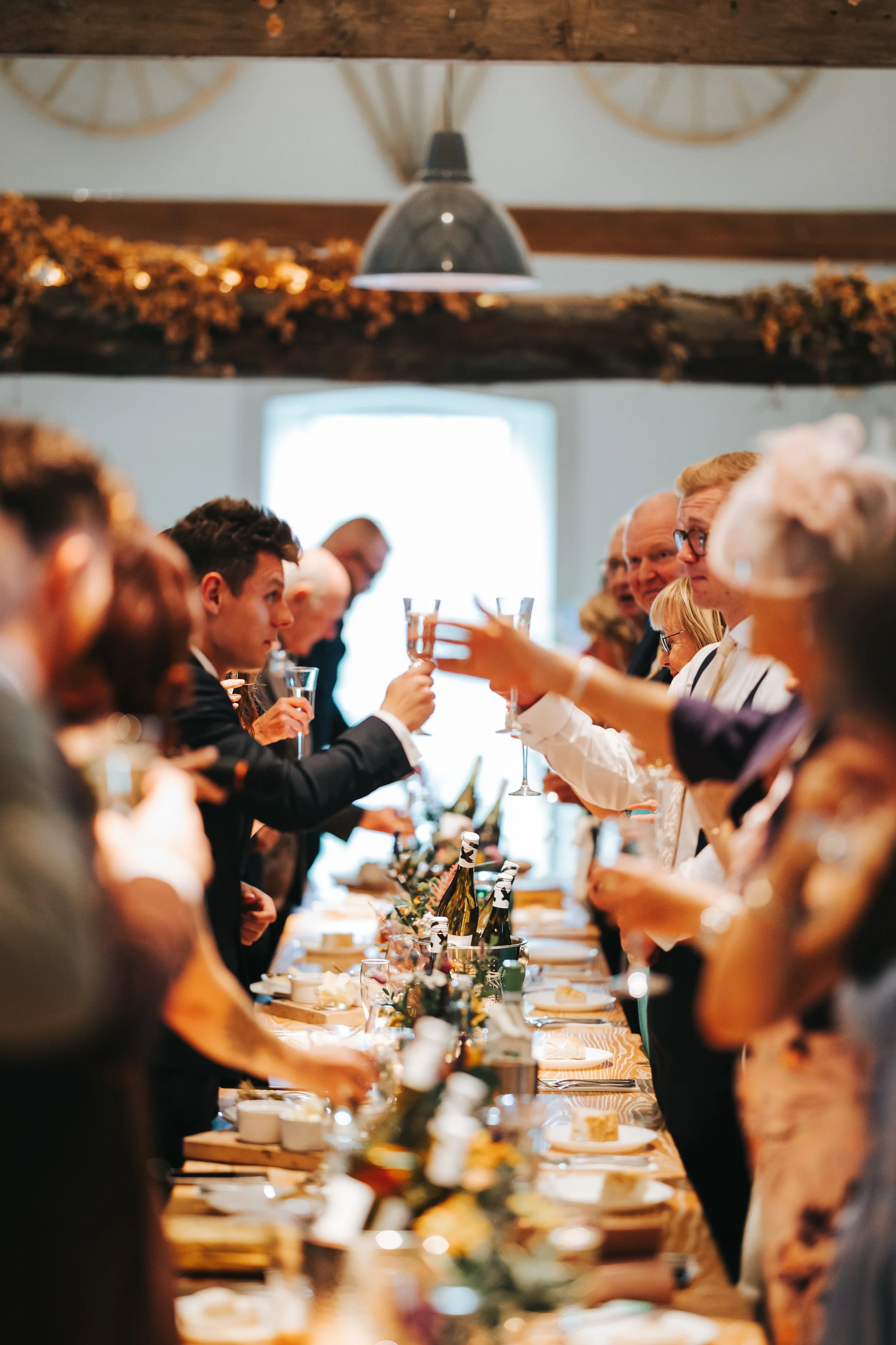 People raising glasses in a toast during a celebration at a long dining table with food and drinks, in a rustic indoor setting with wooden beams and decorative garlands.